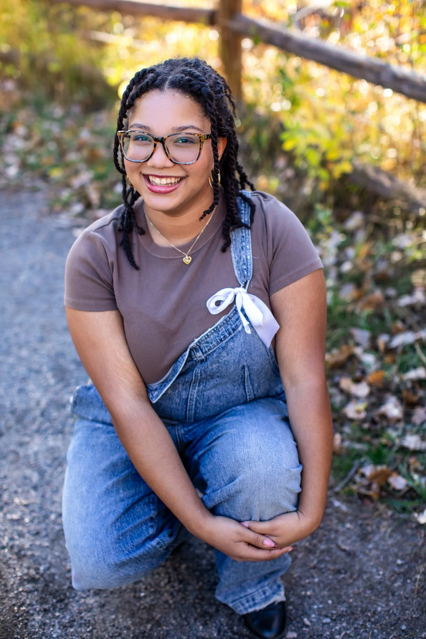 Boulder High School Senior Portrait Photographer gets portrait of young black girl in overalls with a white ribbon holding her overalls up kneeling down on the ground.

