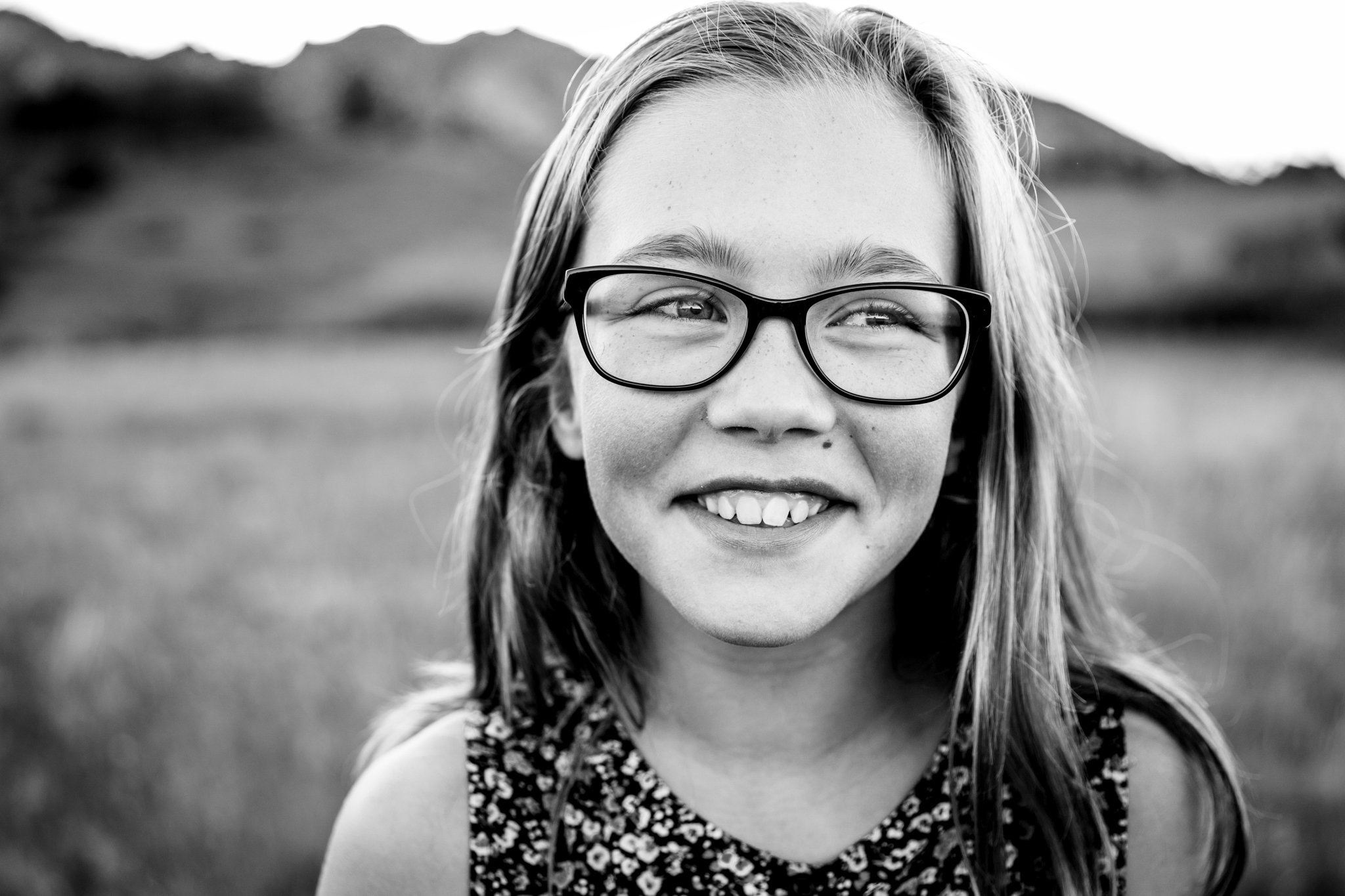 A young girl with glasses smiling outdoors in black and white, with hills in the background.
