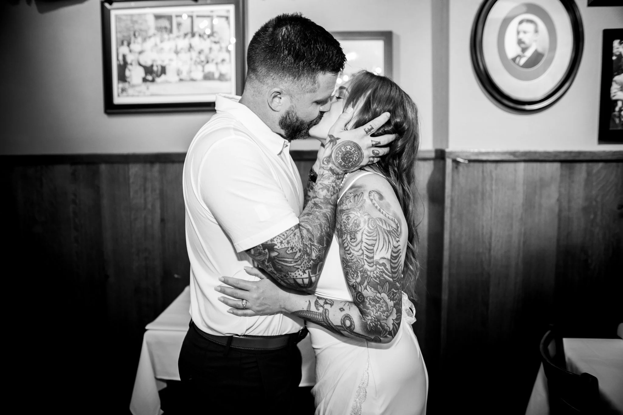 A tattooed man and woman sharing a kiss in a dining room, with framed pictures on the wall in the background.