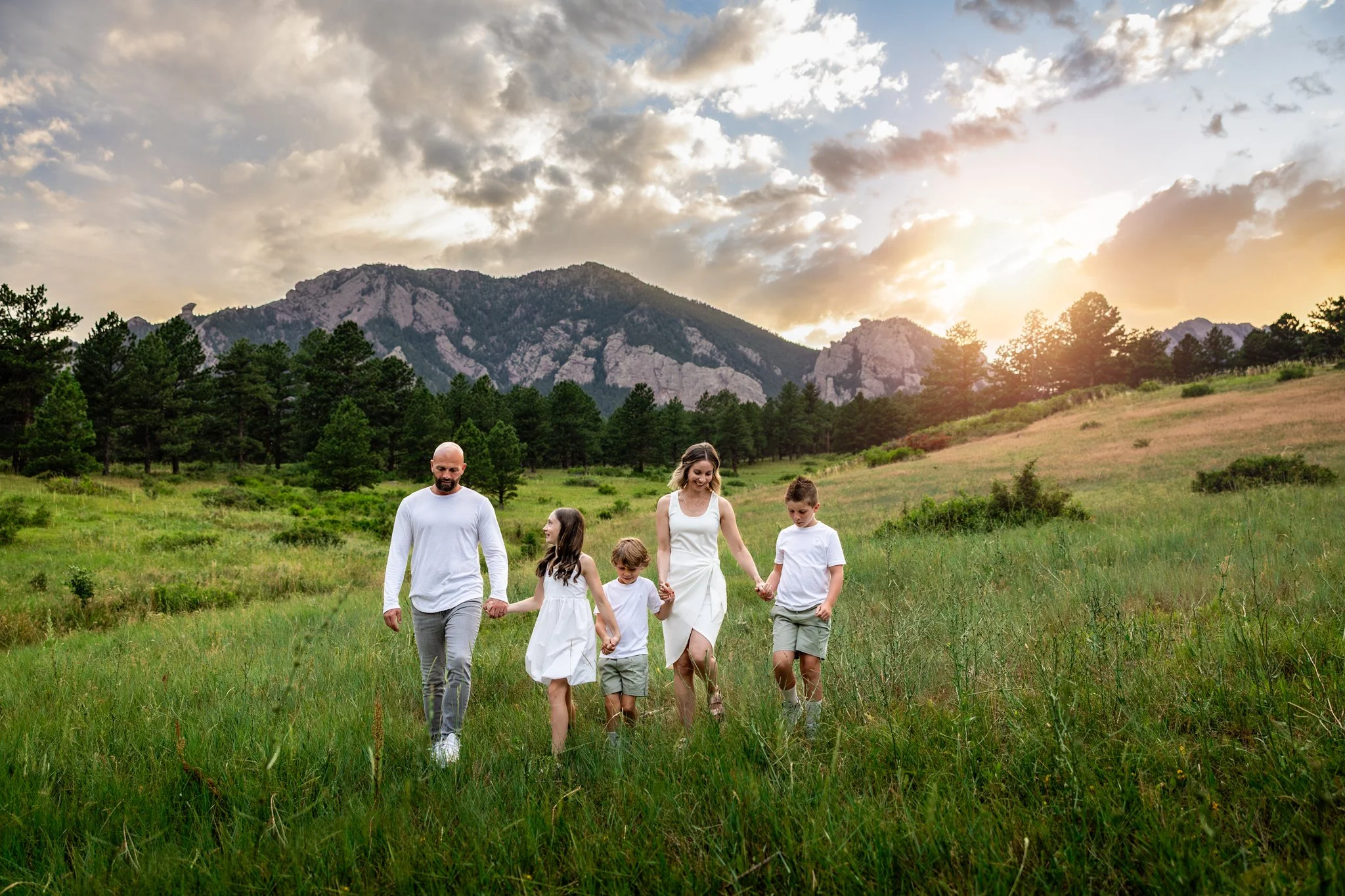 Colorado family photographer takes a portrait of a family walking through a lush green field with the mountains in the background at sunset.