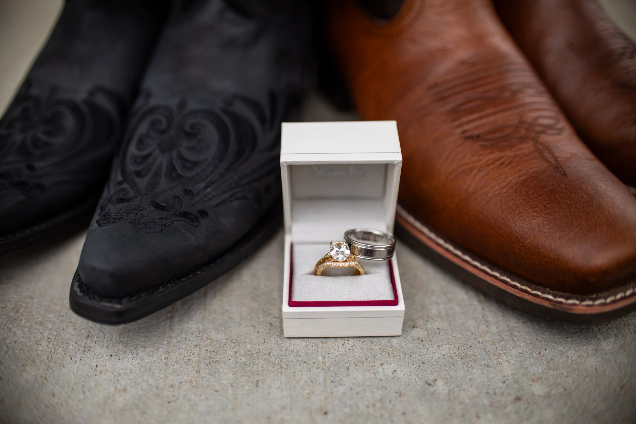 A white jewelry box with pink lining holding two rings, one gold with a large diamond and one silver band, placed on a surface between a black patterned dress shoe and a brown leather dress shoe.