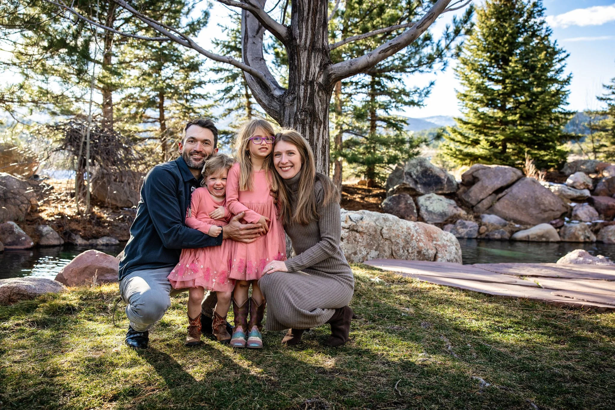 A family of four, including two children and two adults, poses happily outdoors near a tree by a small river in a forested area during daytime.