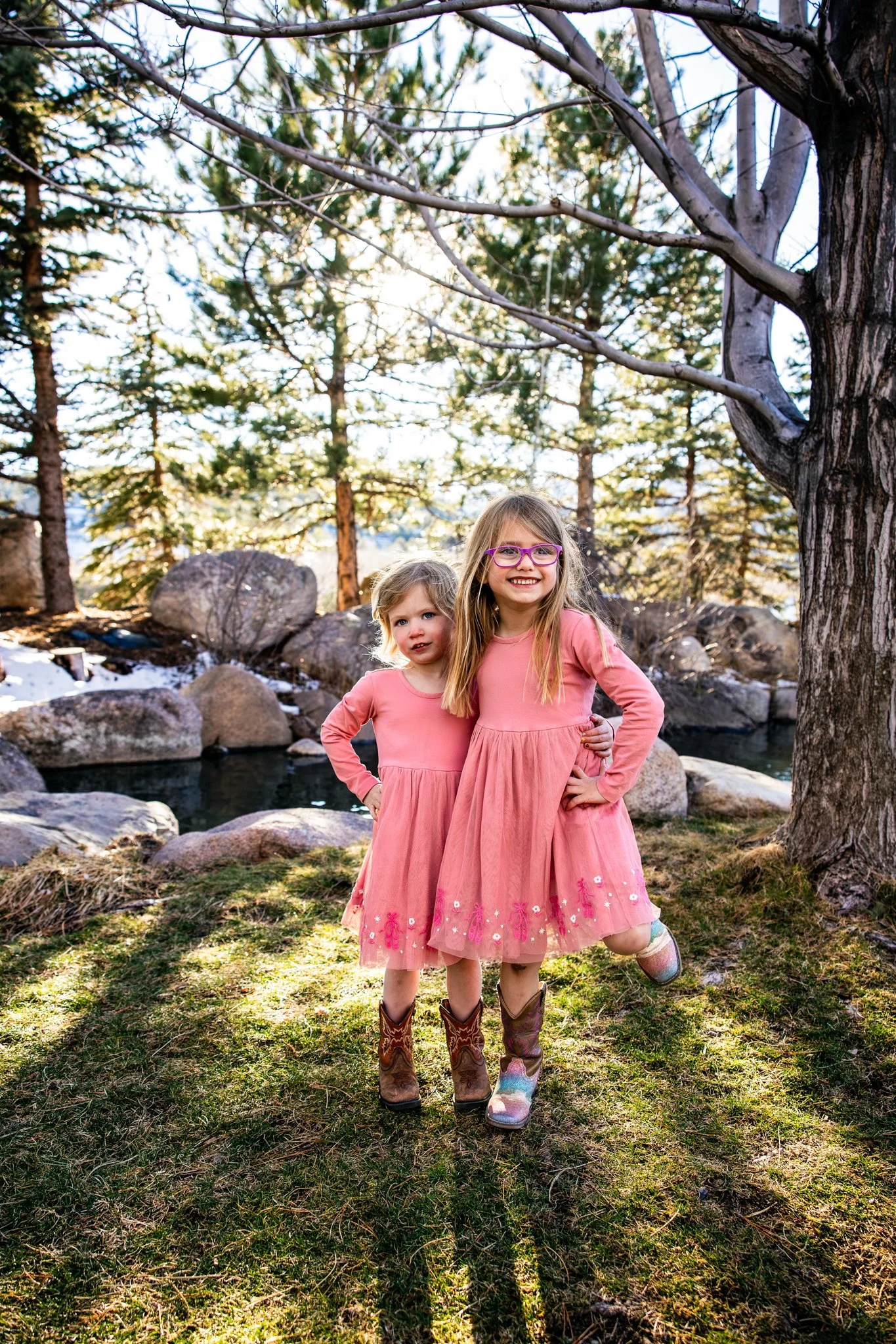 Two young girls in pink dresses and cowboy boots standing outside near a tree with a pond and rocks in the background during daylight.