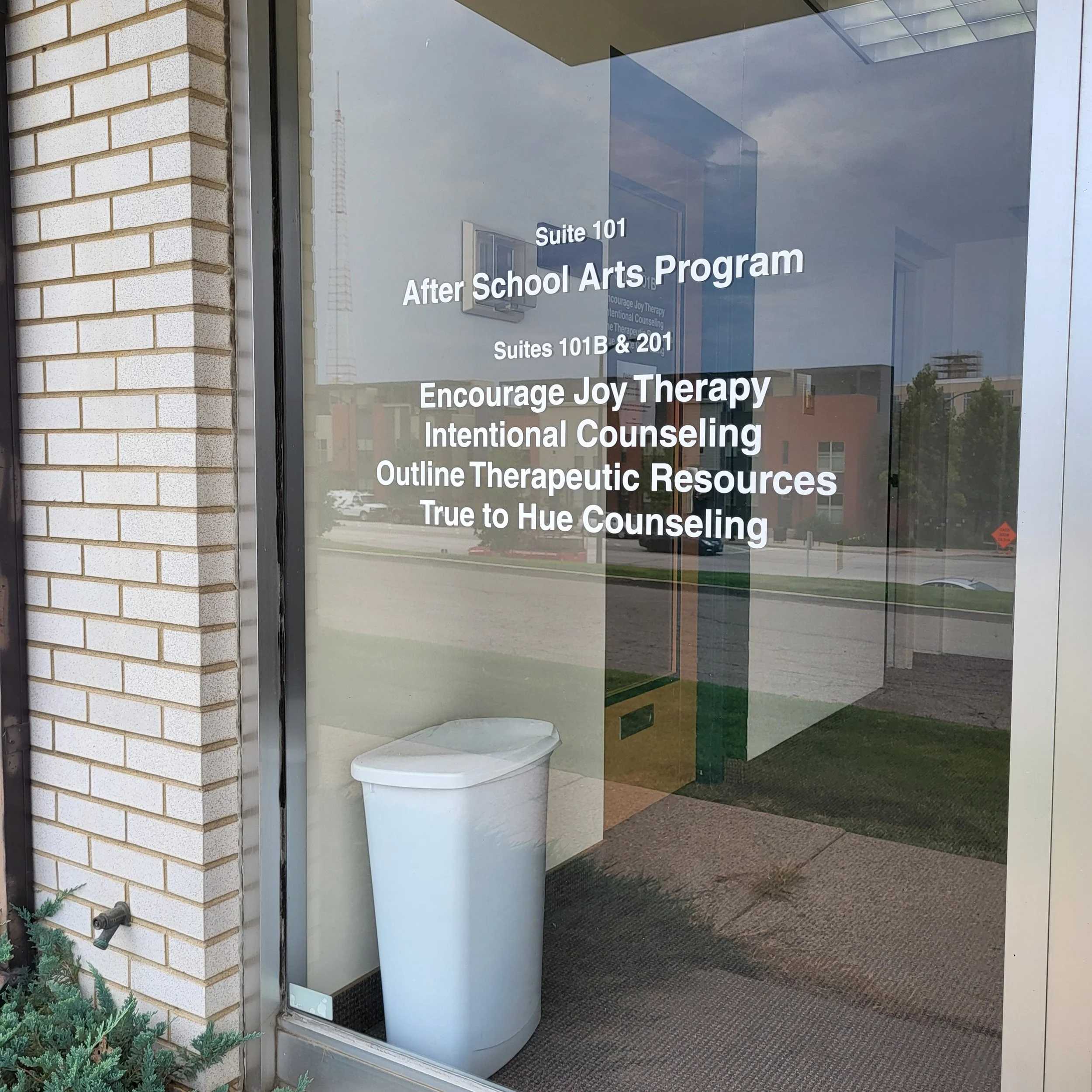 Glass door with white text listing programs and classes, with a white trash can inside, outside reflection of a parking lot and buildings.