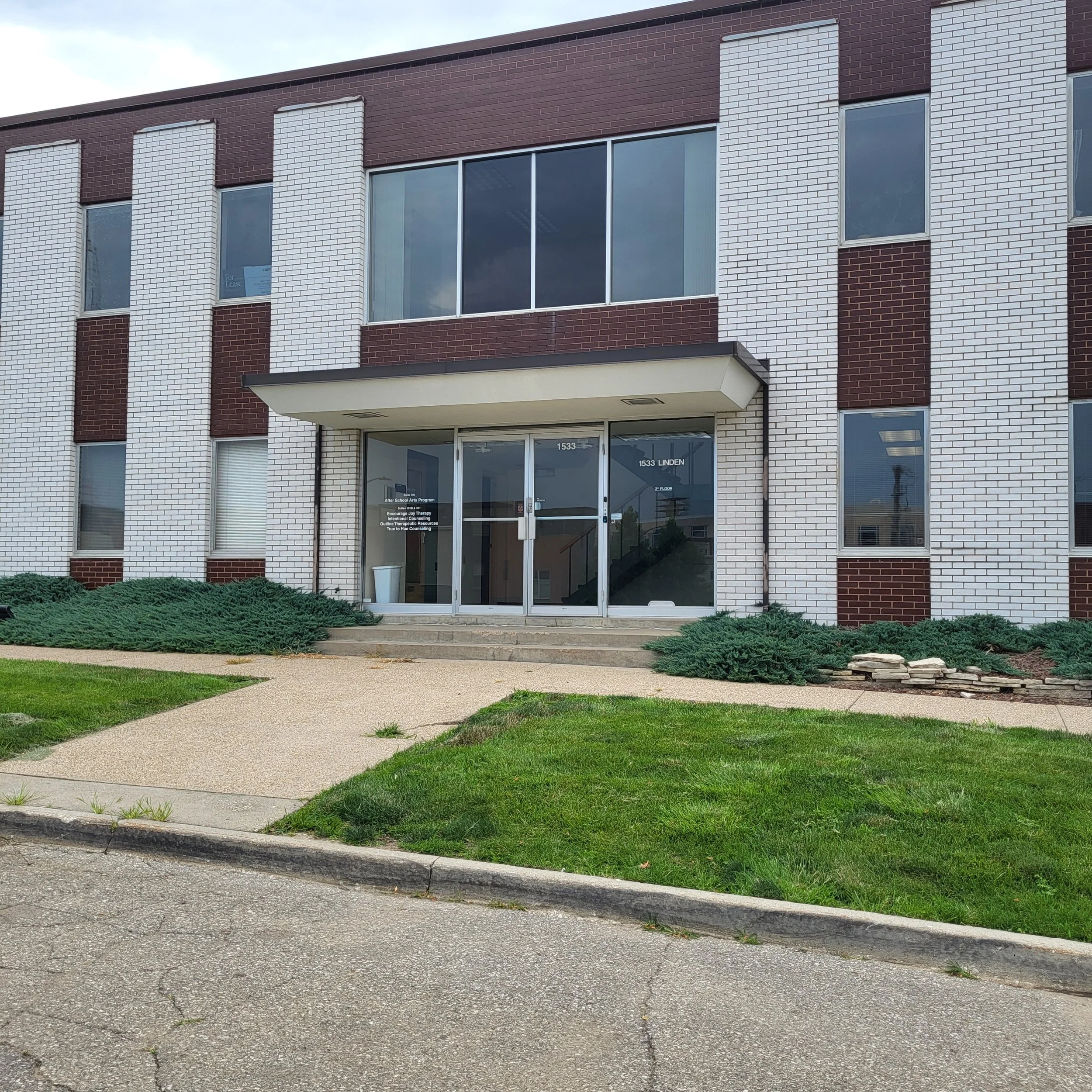 Entrance of a modern brick building with glass doors, stairs leading up to it, and well-kept landscaping including bushes and grass in front.