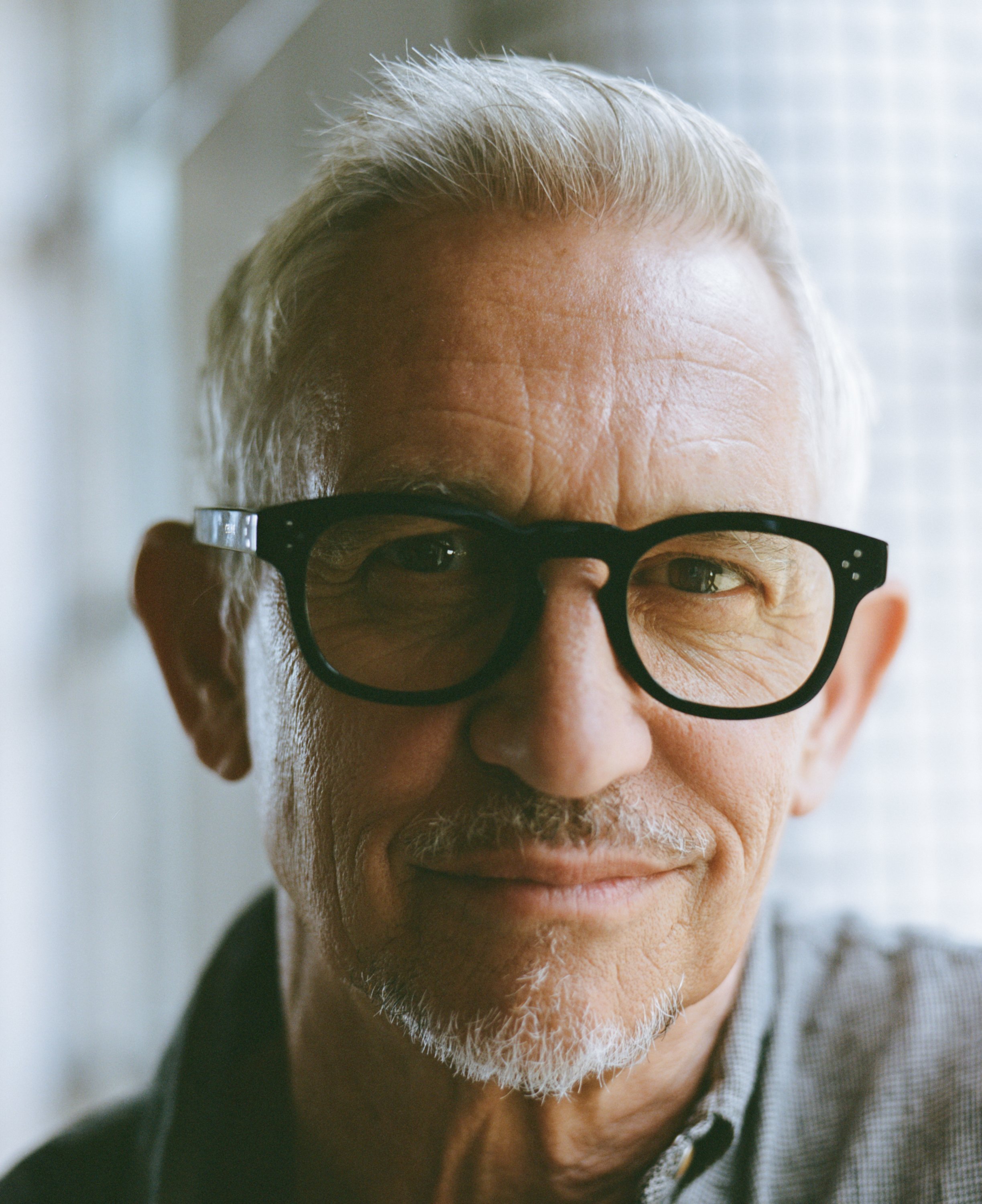 Close-up of an older man with gray hair, glasses, and a mustache, smiling softly, standing near a window.