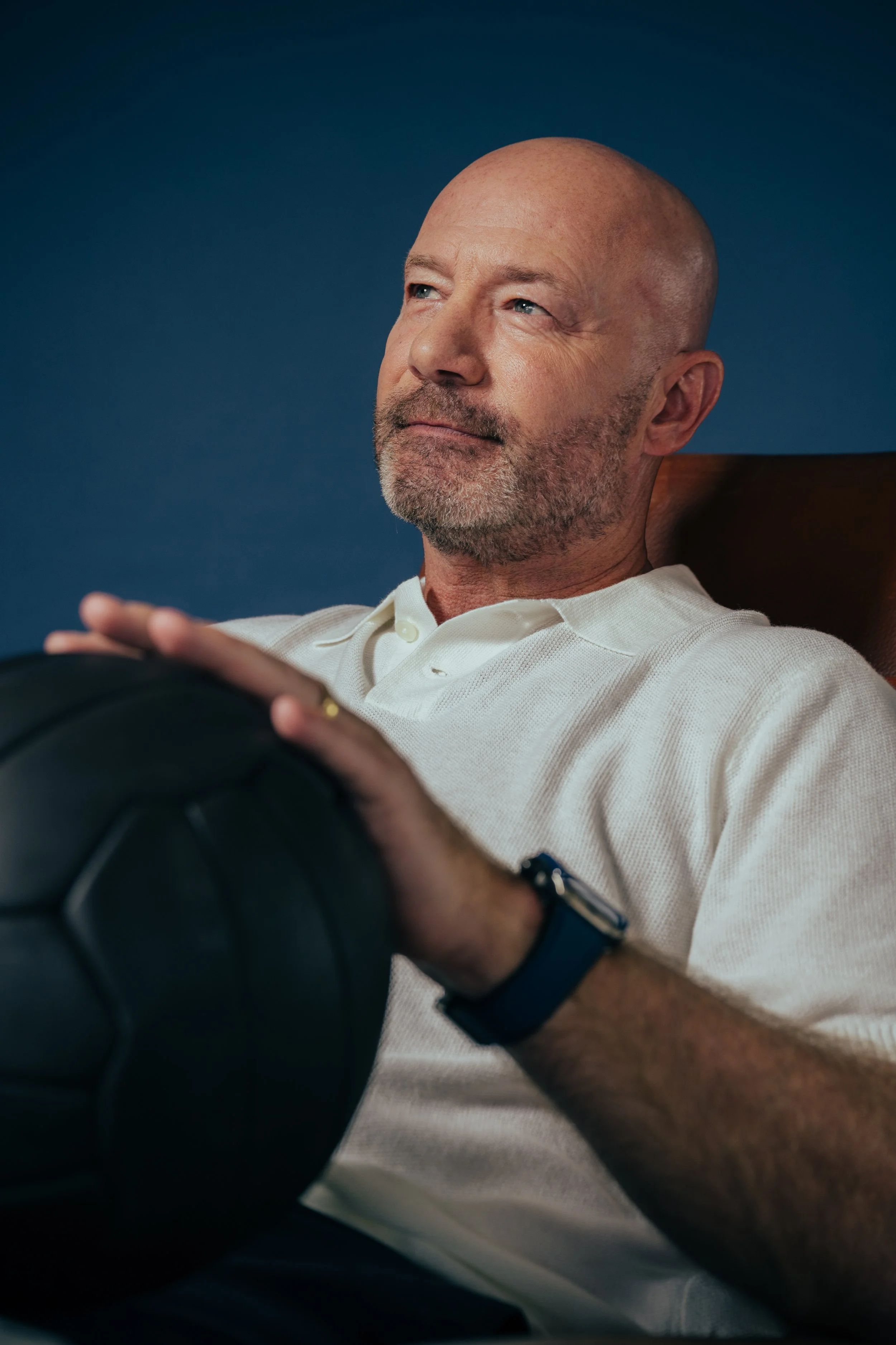 A middle-aged man with a shaved head and beard, sitting in a chair, holding a black football.