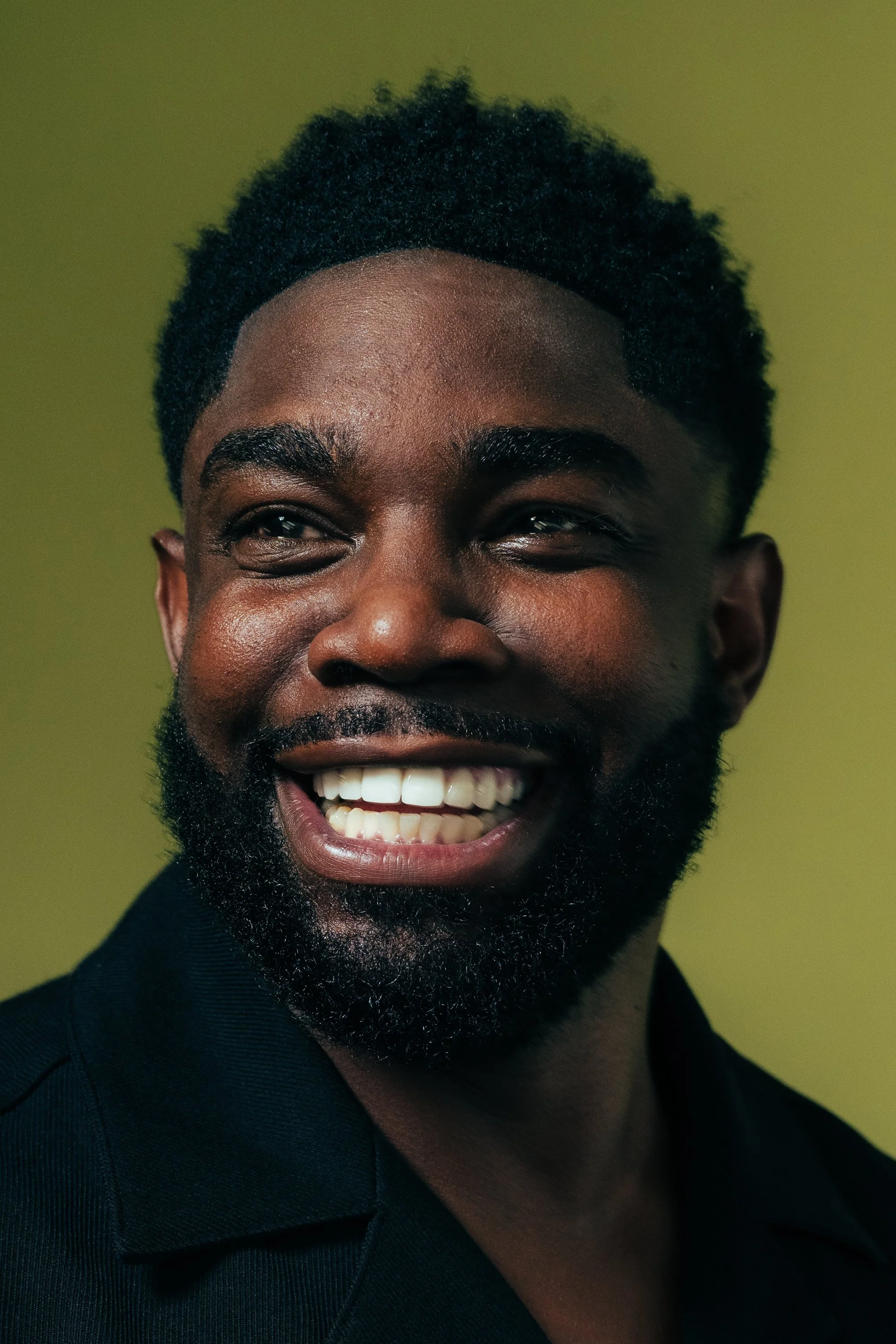 Close-up of a smiling man with a beard and curly hair, looking to the side, against a green background.