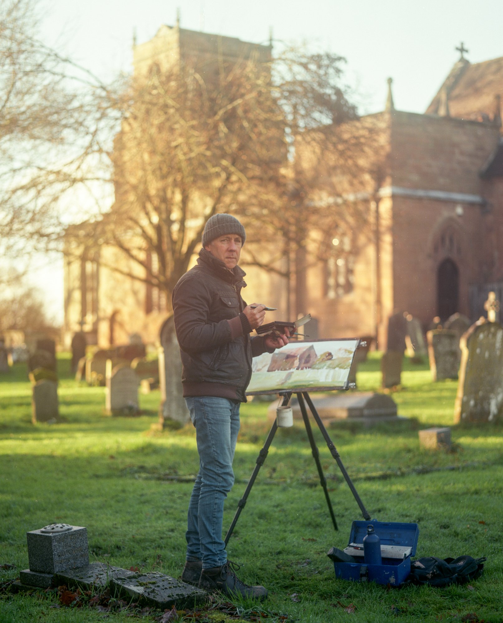 A man standing outdoors in a graveyard, working on a painting on an easel, with a church in the background during sunset.