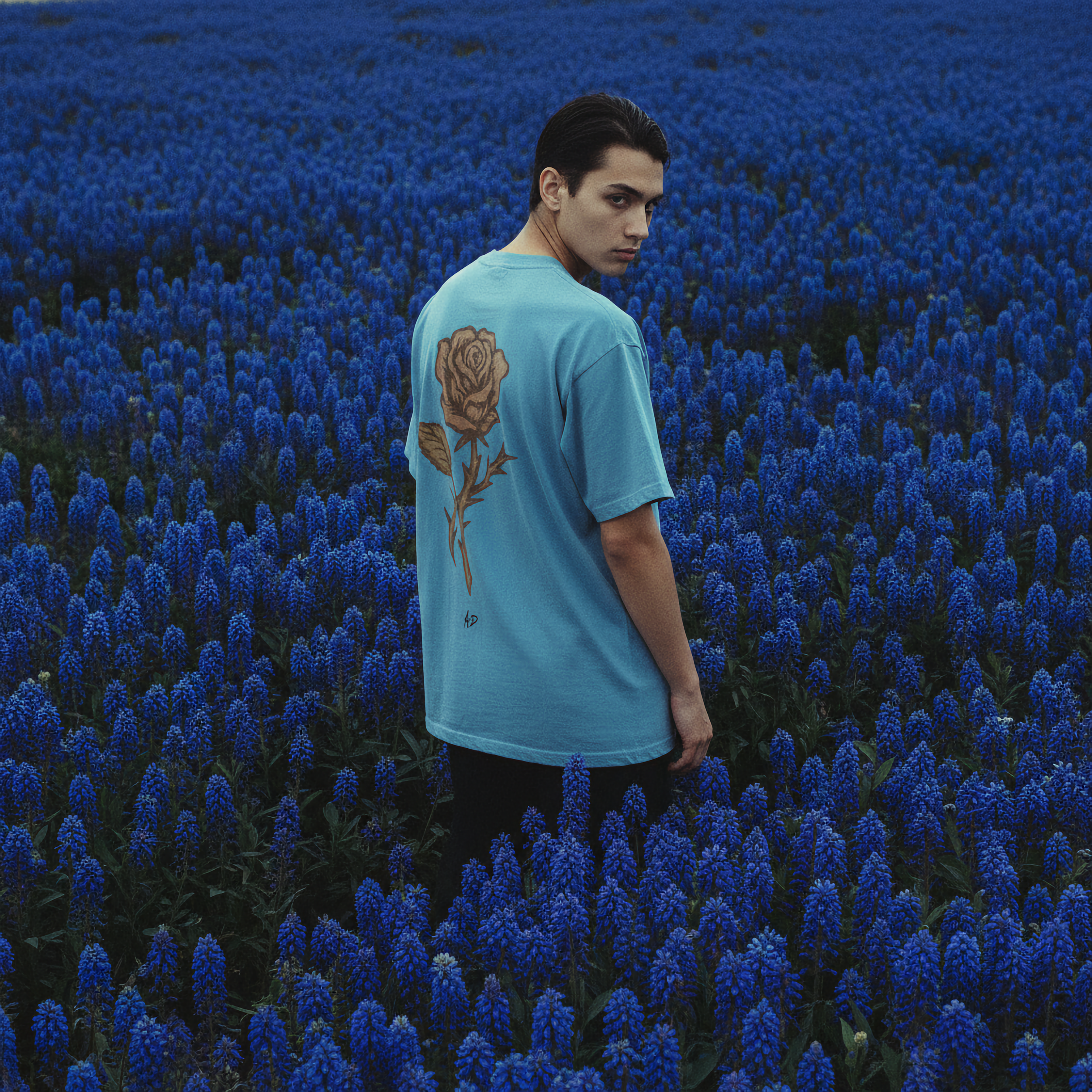 A young man with dark hair and a focused expression standing in a field of vibrant blue flowers, wearing a light blue T-shirt with a floral tattoo design on his shoulder.