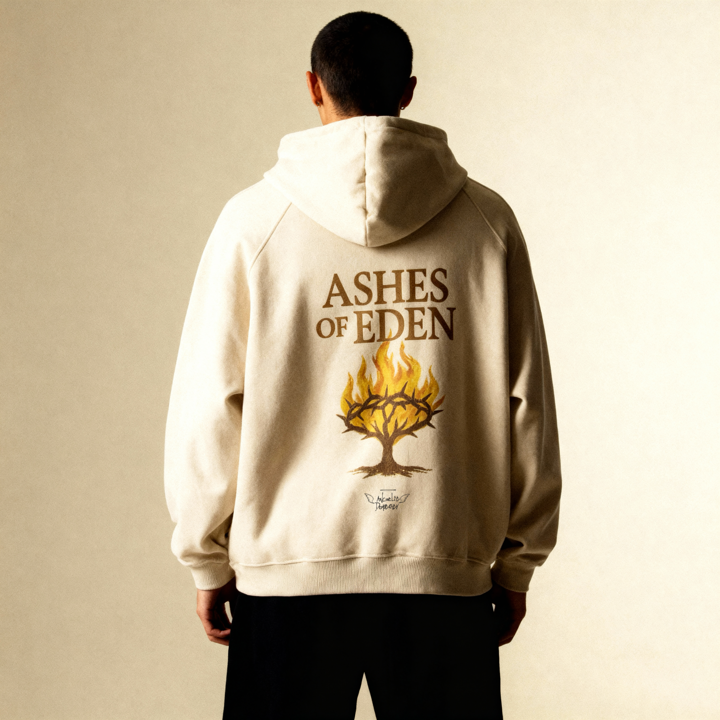 Back view of a person wearing a beige hoodie with the words 'ASHES OF EDEN' and a fiery tree graphic, standing against a beige wall.