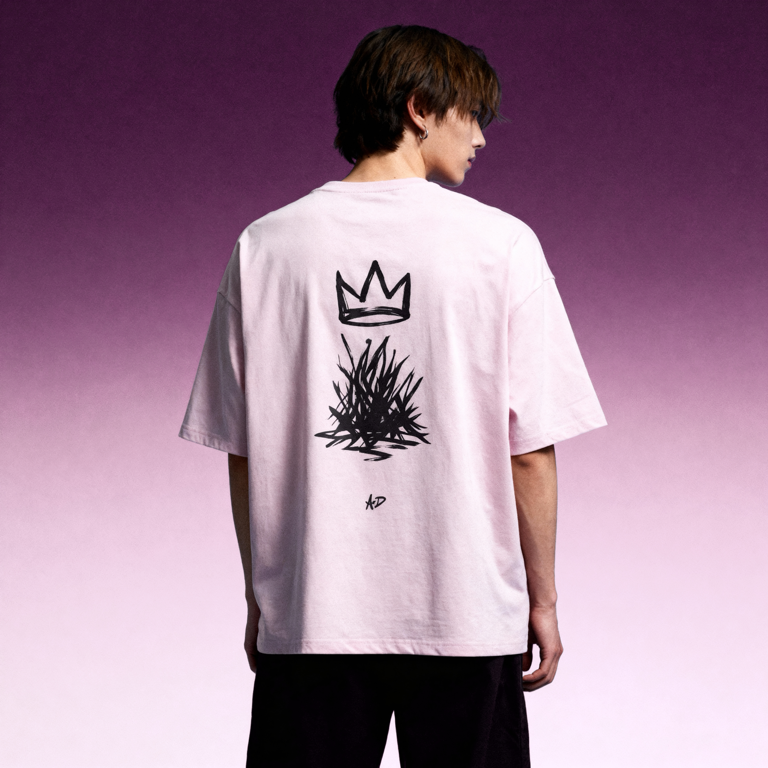 A person with short brown hair and hoop earrings wearing a pink t-shirt with a black crown graphic on the back, standing against a gradient purple background.