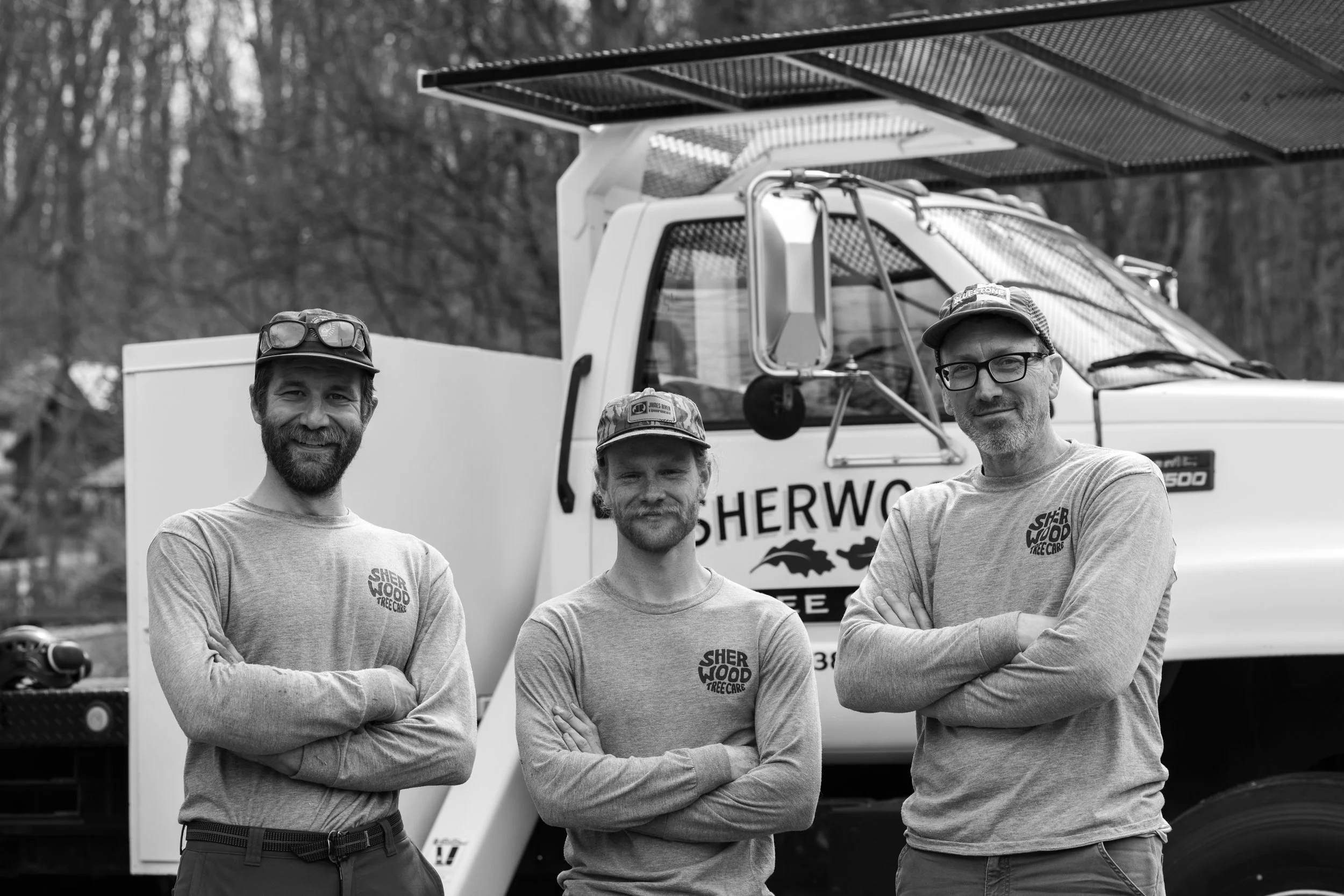 Three men standing in front of a large service vehicle, all wearing long-sleeve t-shirts with the Sherwood Tree Care logo, smiling and crossing their arms.