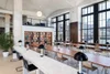 Modern office space with large windows, long tables with black lamps, and a bookshelf with decorative items and books.