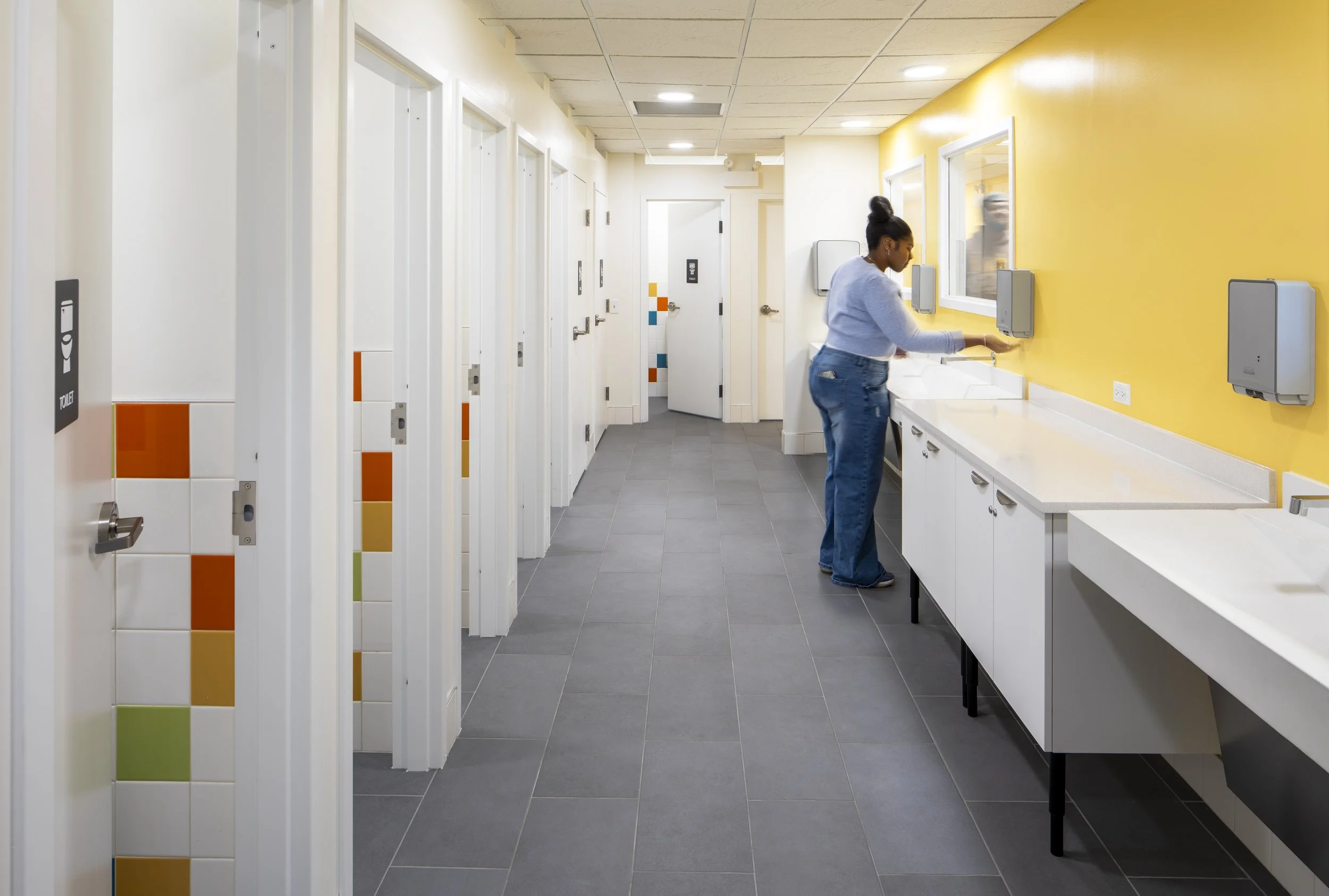 Accessible public restroom with a toilet, grab bars, a paper towel dispenser, a trash bin, a yellow wall, and colorful tiles on the wall.