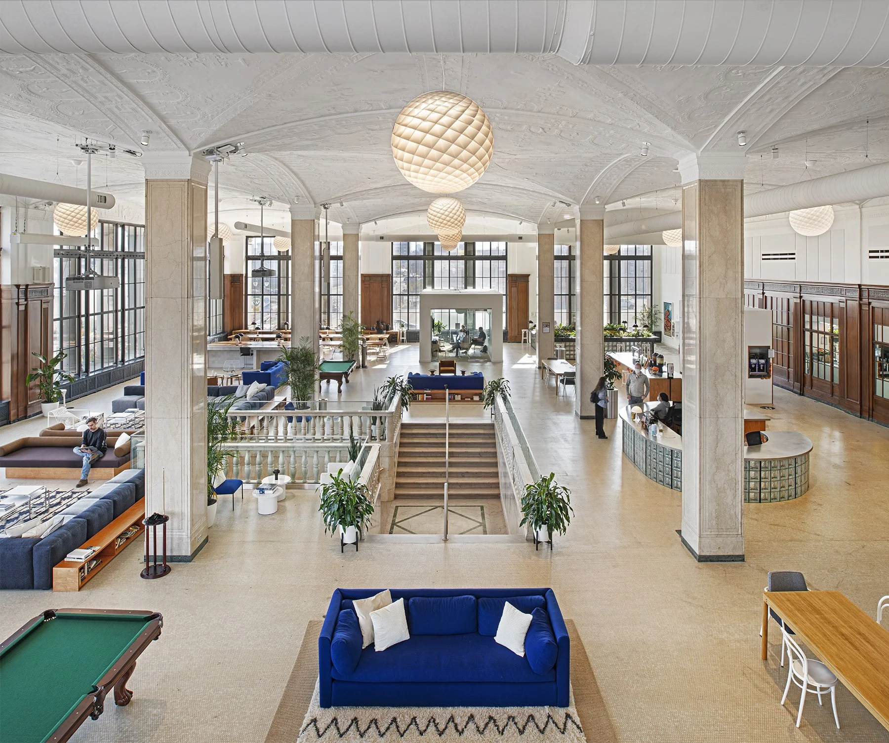 A large beige stone building with multiple floors and numerous window panes, located on a city street with parked cars in front, under a bright blue sky with some clouds and tree branches overhead.