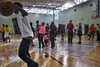 People gathered in a gymnasium, some engaging in conversations, others observing photos displayed on boards, with a person holding a basketball in the foreground.