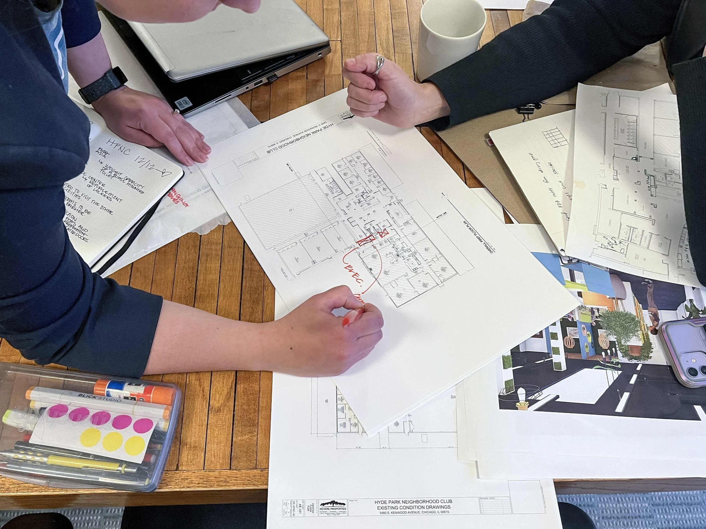 Two people reviewing architectural plans and drawings on a wooden table, surrounded by notebooks, pens, markers, and a laptop.