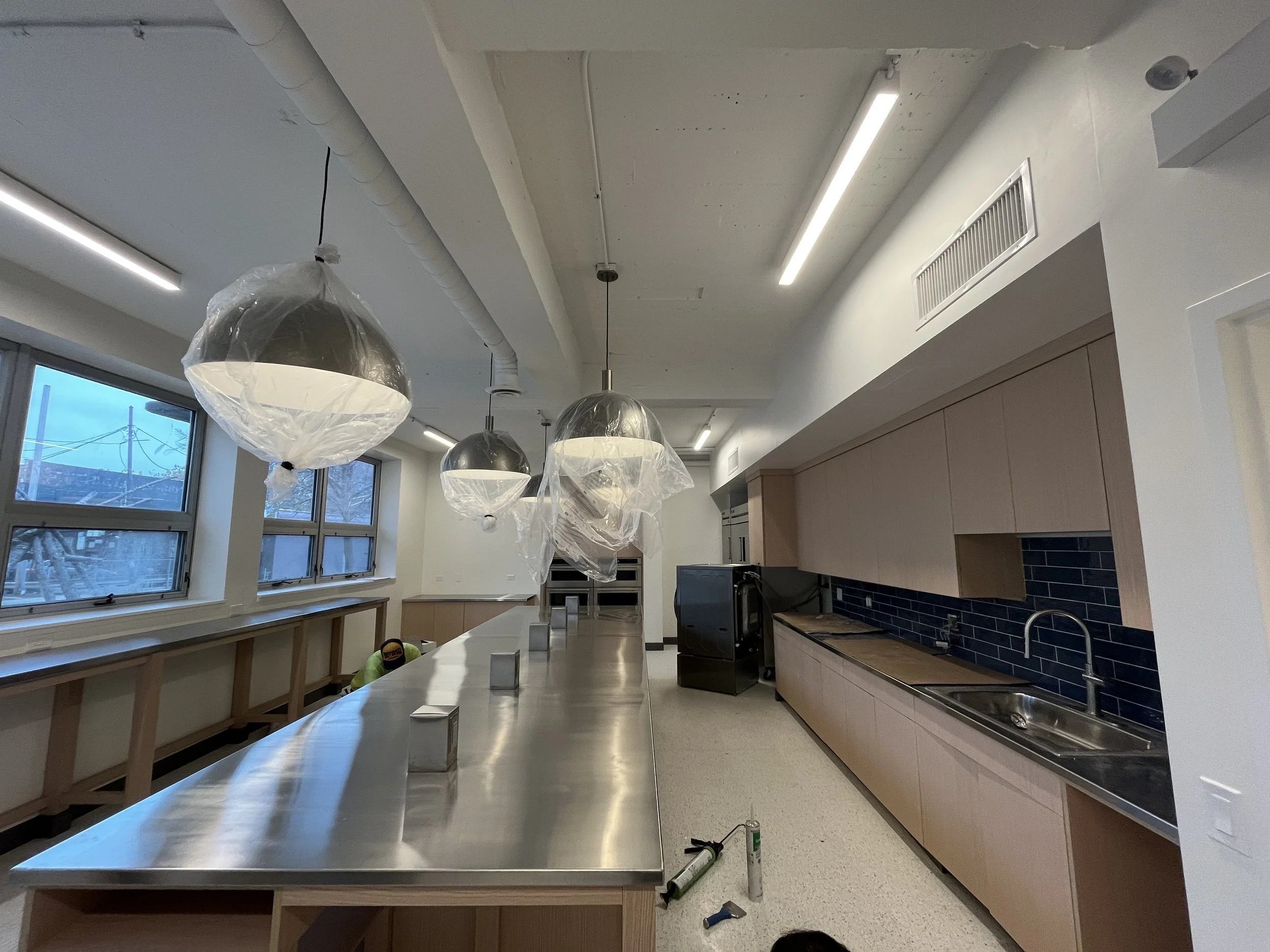 Newly renovated kitchen with wooden cabinets, black tile backsplash, stainless steel sink, and large windows. Three pendant lights are covered in plastic, indicating ongoing installation or cleaning.