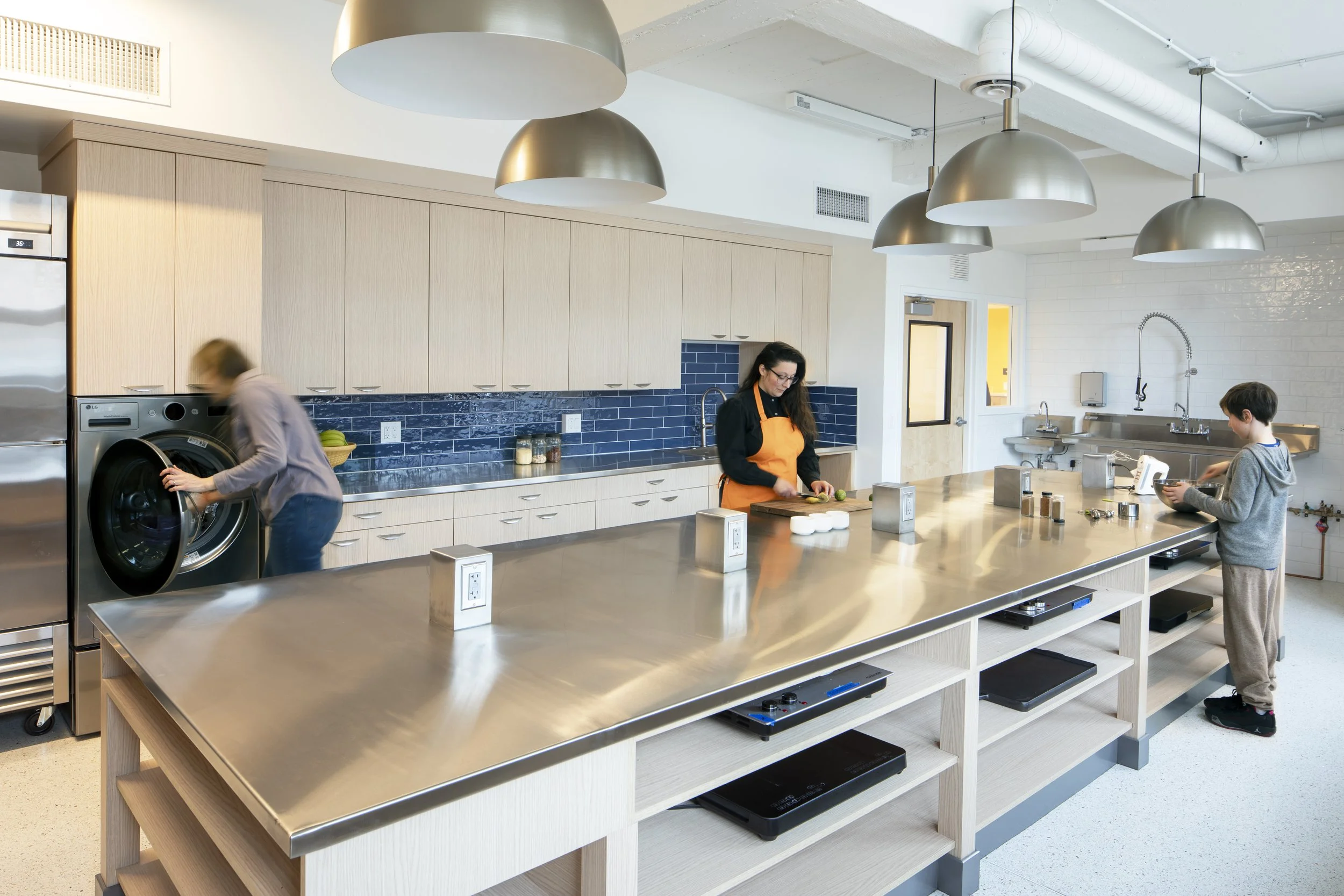 Newly renovated kitchen with wooden cabinets, black tile backsplash, stainless steel sink, and large windows. Three pendant lights are covered in plastic, indicating ongoing installation or cleaning.