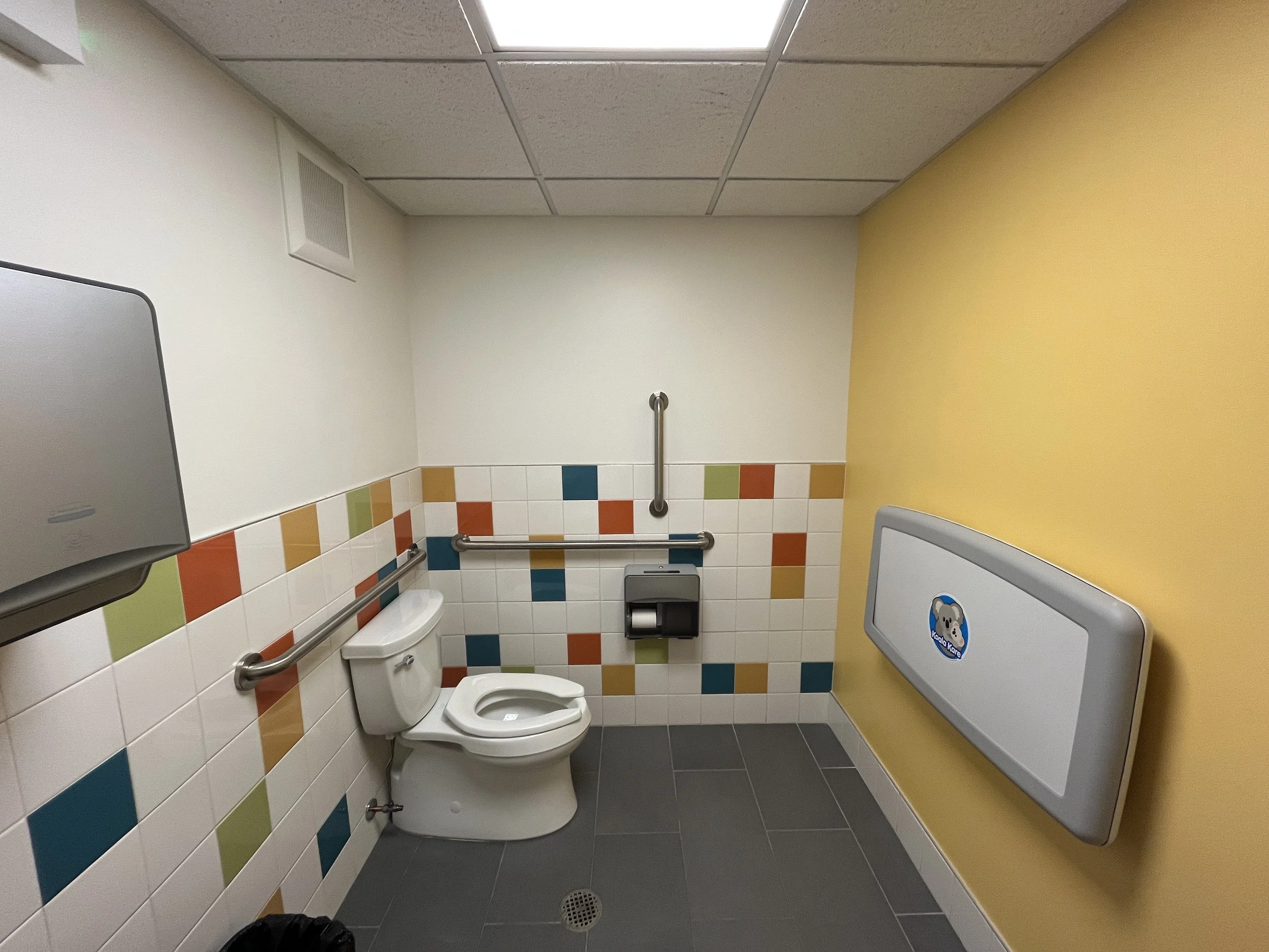 Accessible public restroom with a toilet, grab bars, a paper towel dispenser, a trash bin, a yellow wall, and colorful tiles on the wall.