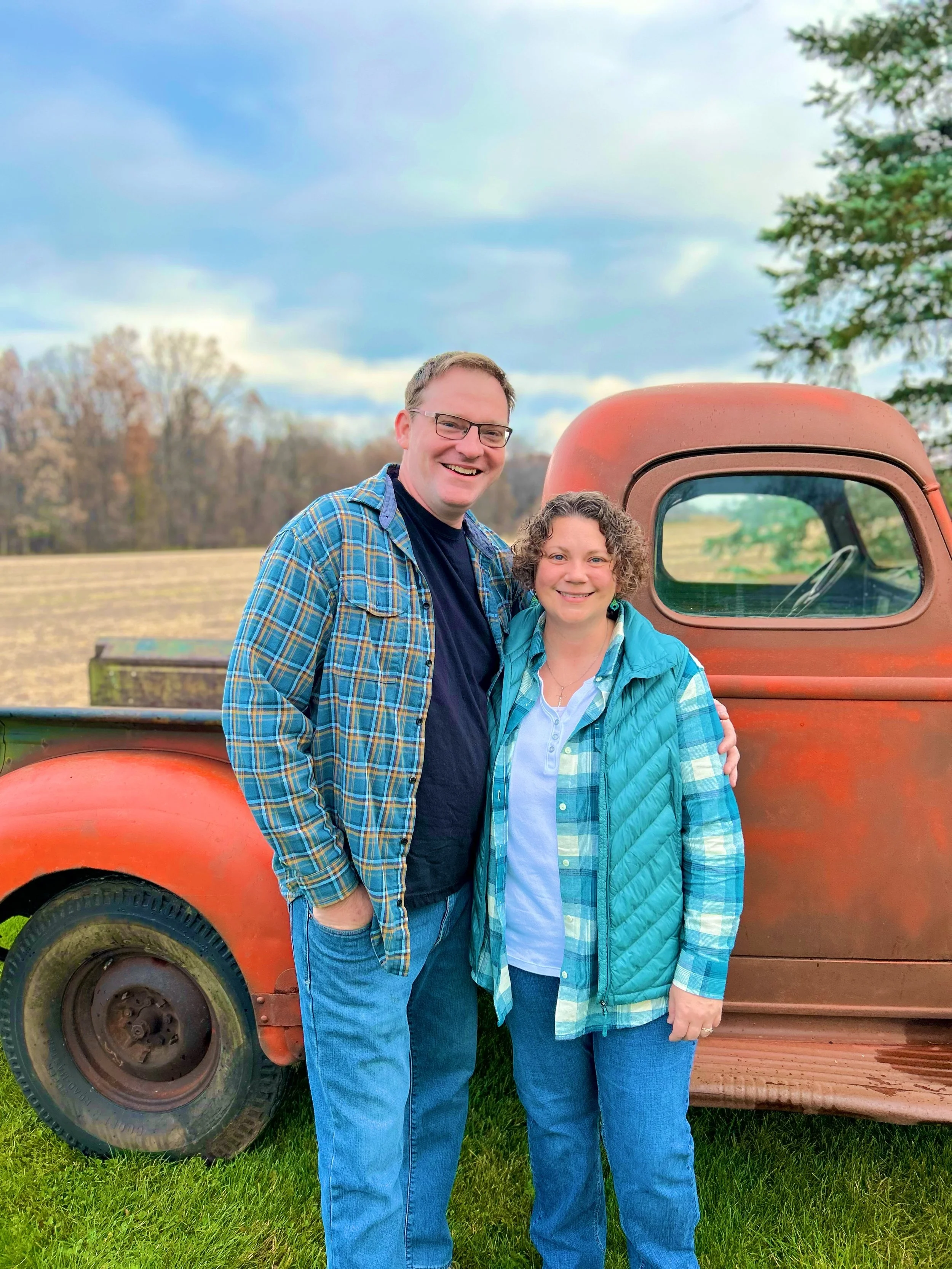 A man and woman standing close together, smiling, with their arms around each other in front of a vintage red pickup truck, outdoors in a rural setting with grass and trees in the background.