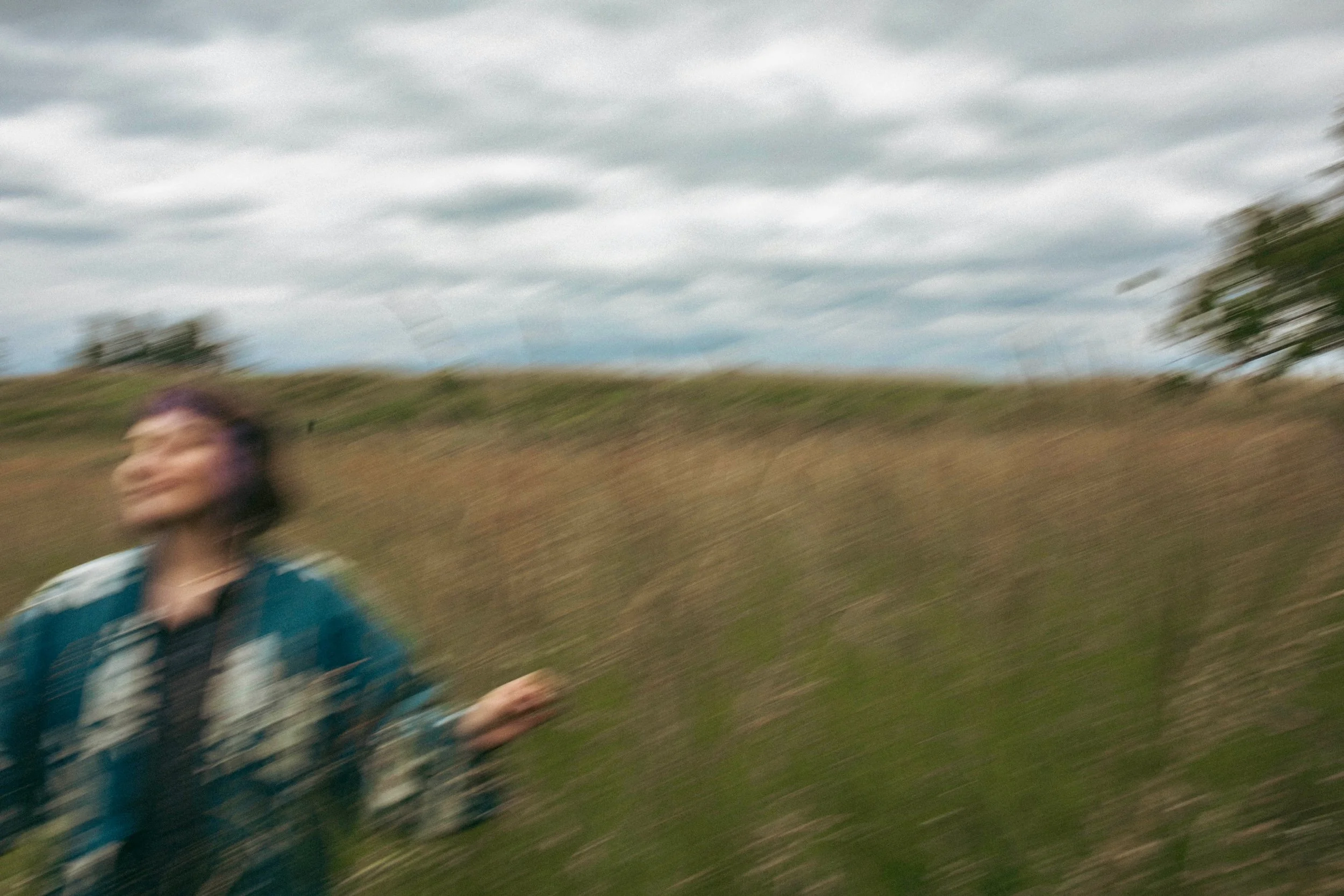 A blurred person standing outdoors in a field with grass and clouds in the sky.