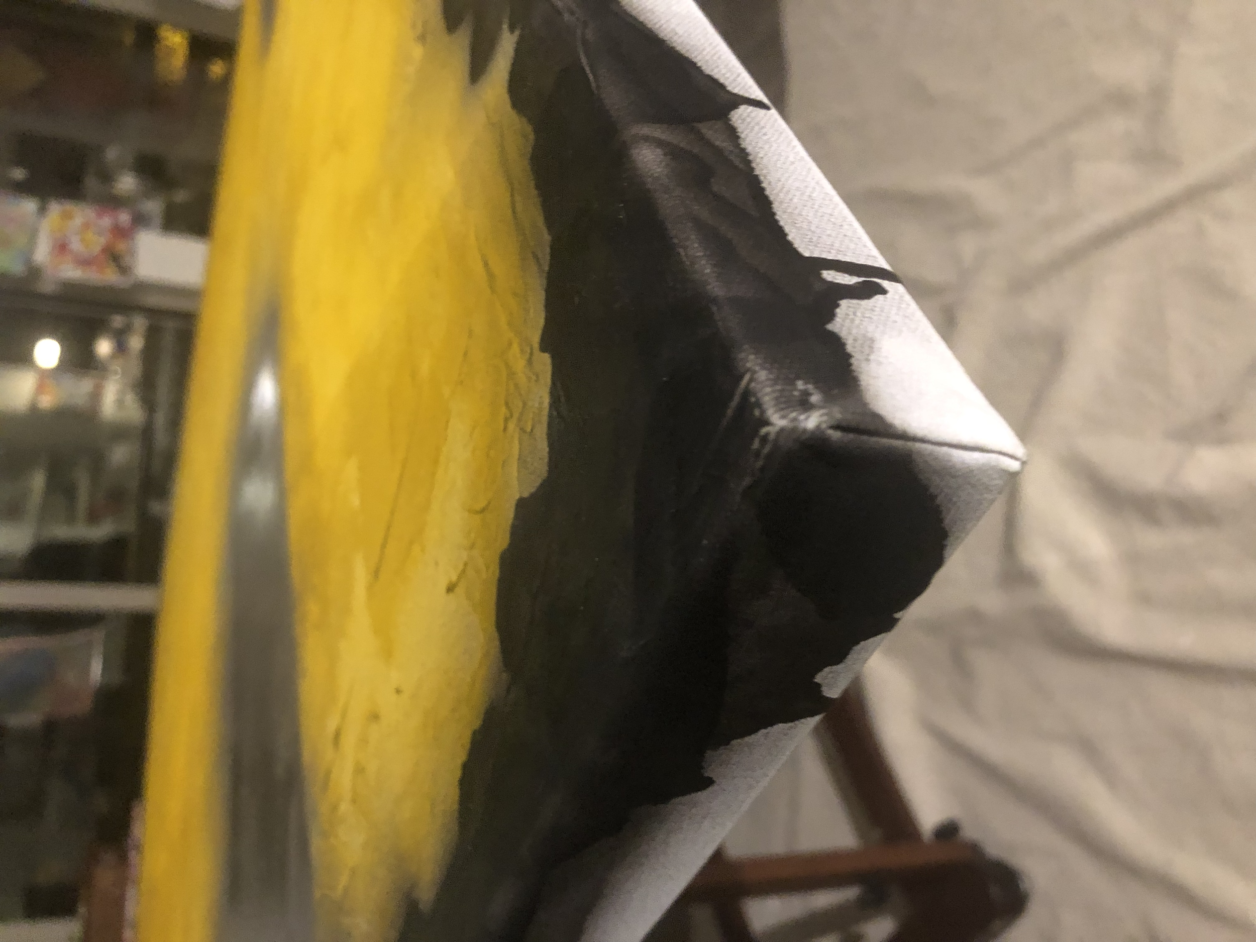 Close-up of a black and white patterned object, possibly a piece of fabric or clothing, on a yellow surface with a background of a room with shelves and various items.