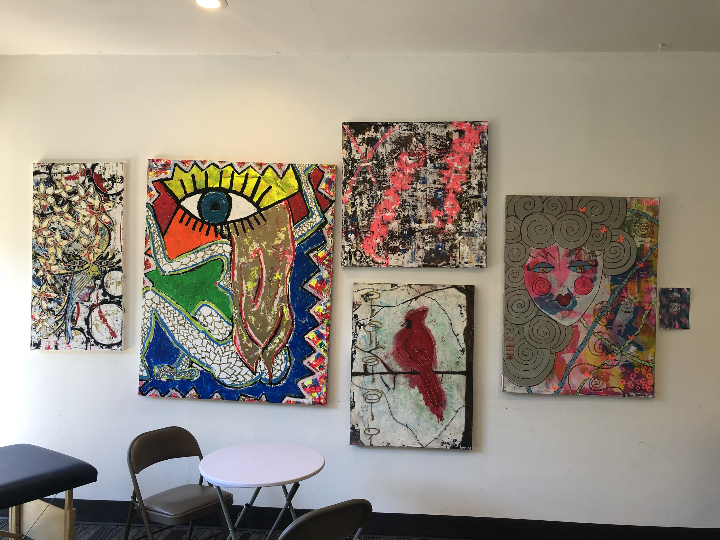 Gallery wall with six colorful abstract paintings of varying sizes, including a large one with a big eye and face, a bird on a branch, and a face with swirling hair, in an art studio or gallery setting.