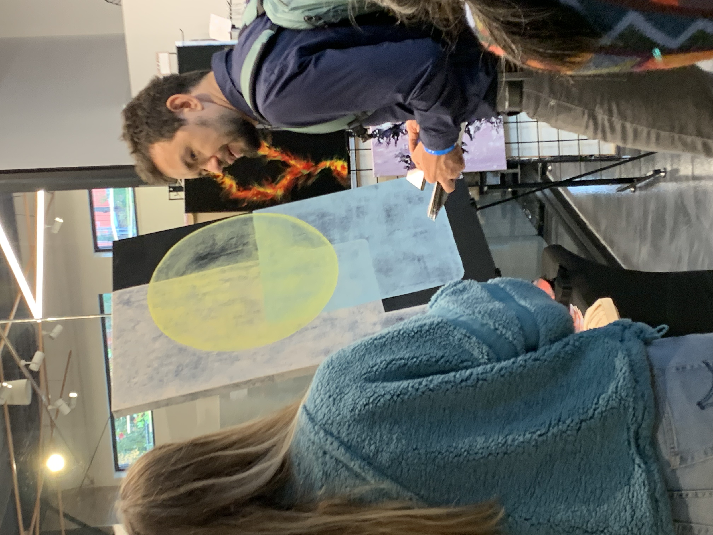Two people, a man with dark hair and a woman with long blonde hair, engaging in conversation in an art gallery. The man is wearing a navy jacket and the woman is wearing a teal fuzzy jacket. They are looking at a piece of art that features a large ye