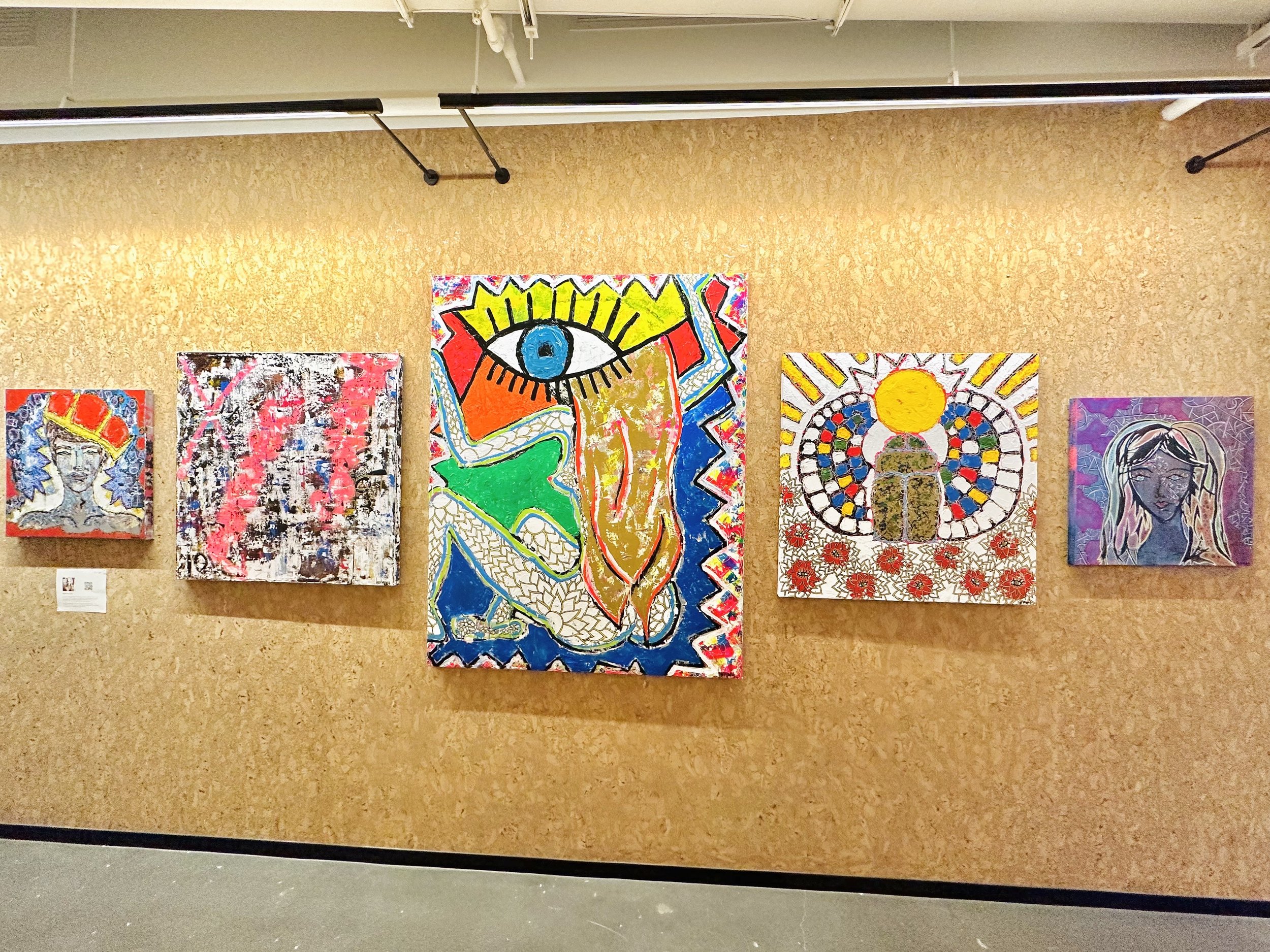 An art gallery wall featuring five colorful paintings, including a portrait of a woman with blue hair, an abstract piece, a large vibrant artwork with an eye and abstract shapes, a mandala with a heart and sun, and a portrait of a woman with long hai