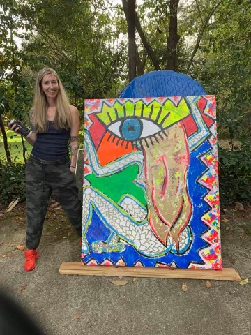 A woman with long blonde hair, wearing a black tank top, dark camouflage pants, and red shoes, standing outdoors next to a colorful abstract painting on an easel. The painting features a large eye, mouth, and various bright shapes, with trees and gre