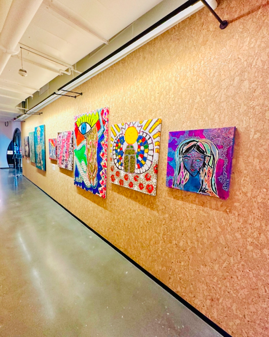 Gallery wall displaying colorful abstract and portrait paintings in an art exhibition space.