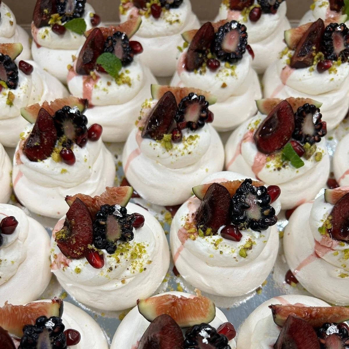 A tray of mini pavlovas decorated with chantilly whipped cream, blackberries, cherries, chopped figs, pomegranate seeds, pistachios, and a small mint leaf.