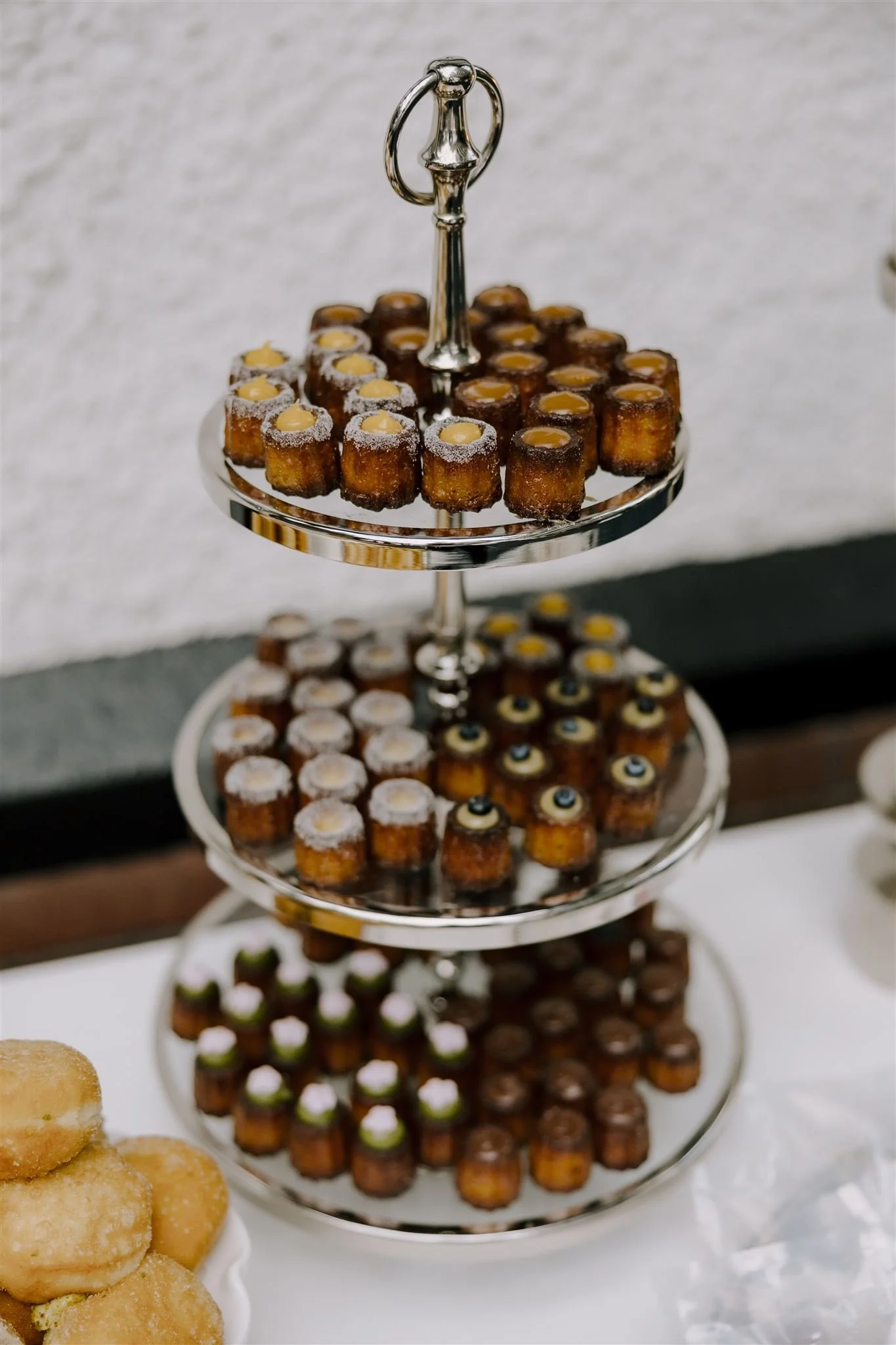 Three-tiered silver dessert stand filled with small baked canelé pastries with decorative toppings, set on a white table, with a white textured wall in the background.