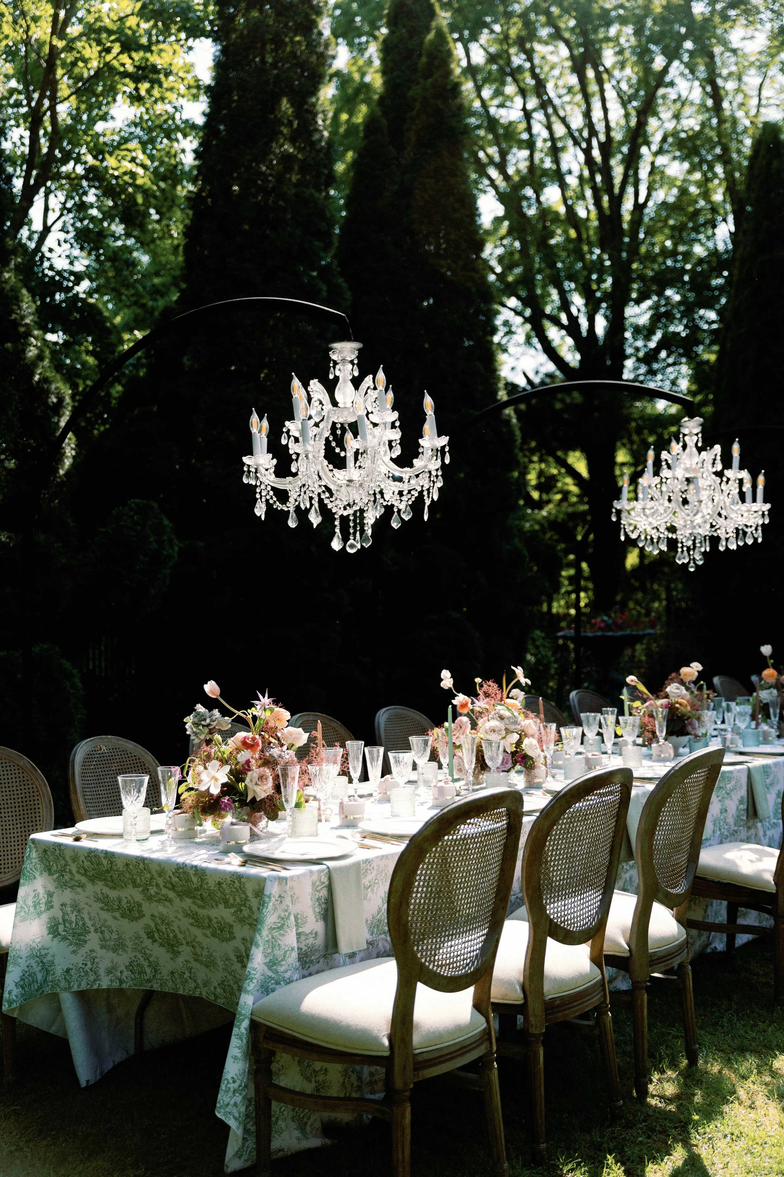 Elegant outdoor dining table with floral arrangements and crystal chandeliers hanging overhead in a lush green garden setting.