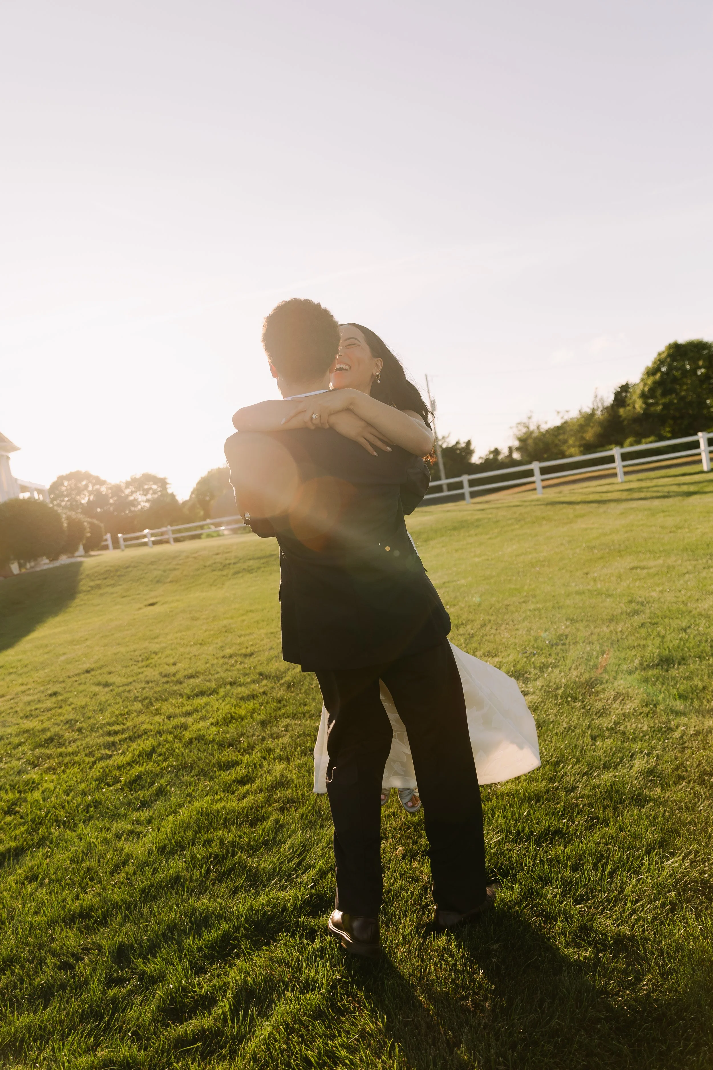 A couple embracing and smiling on a grassy field during sunset, with a white fence and trees in the background.