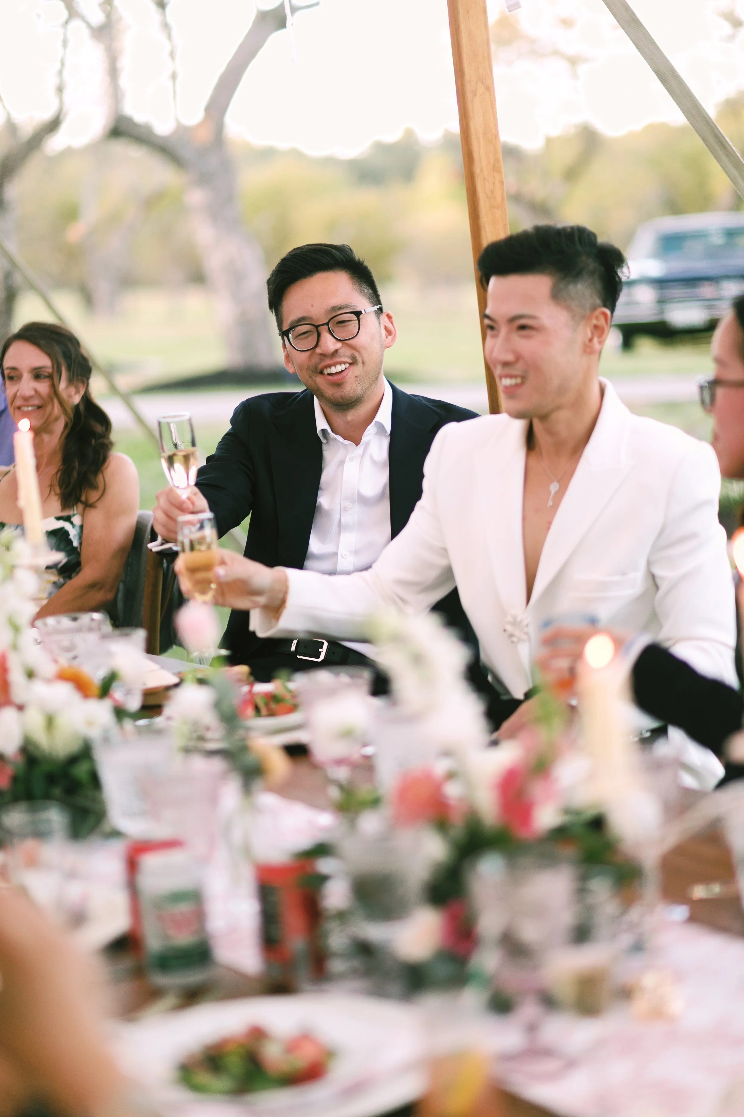 People celebrating at a wedding reception, raising champagne glasses and smiling, with floral arrangements and food on the table.