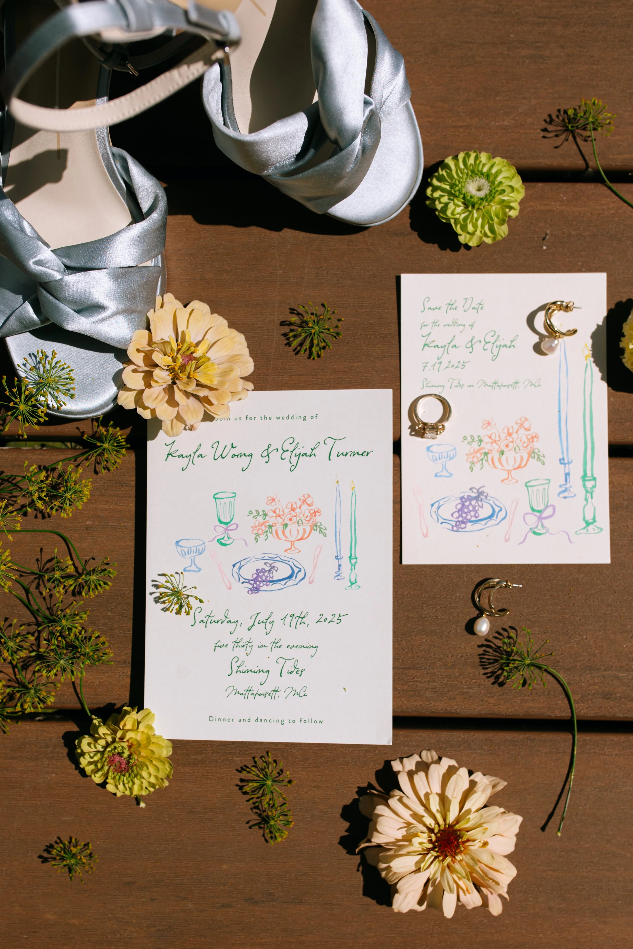 Wedding invitation, jewelry, and flowers on a wooden surface, with light blue satin shoes in the background.