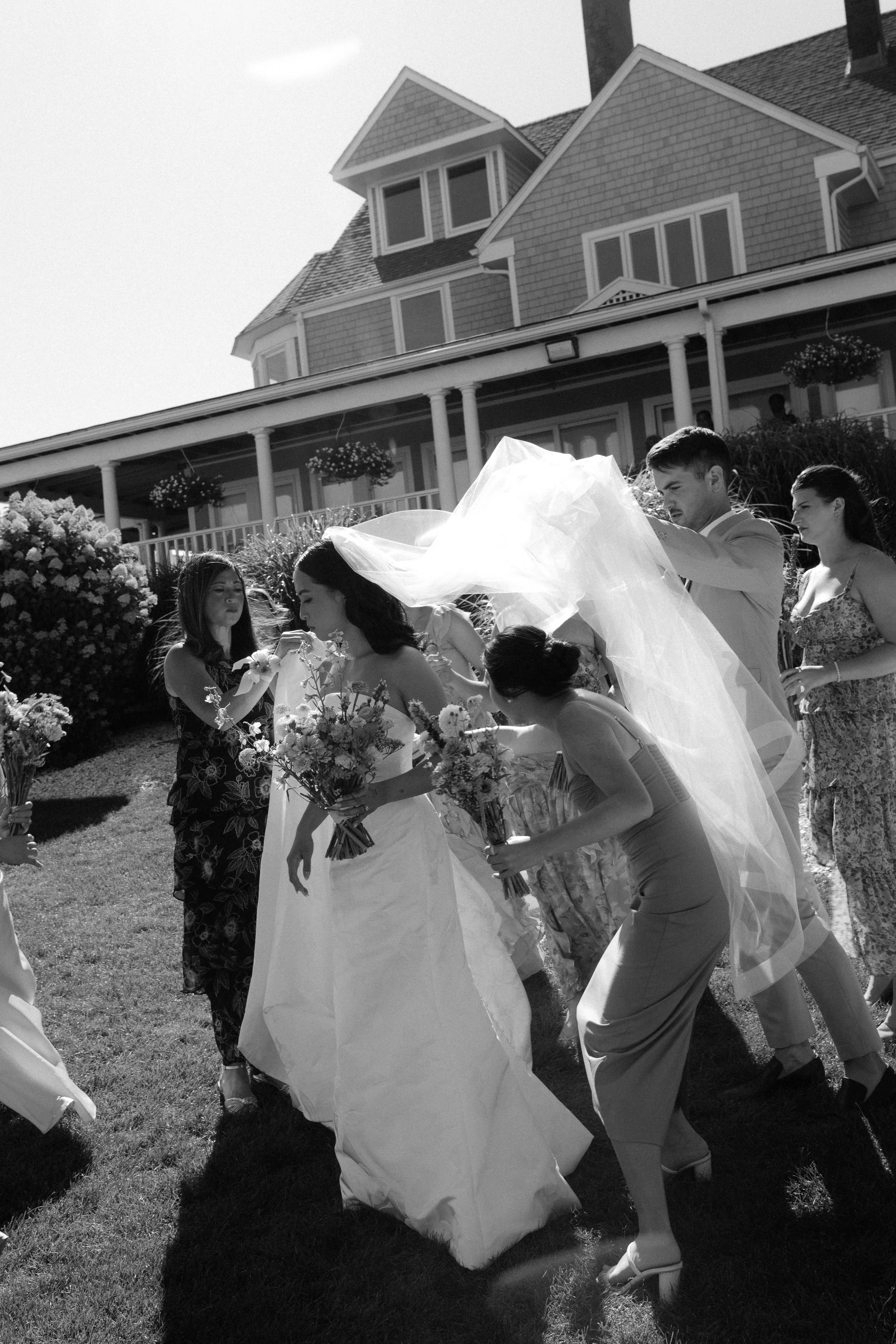 A wedding scene with a bride and entourage outside a large house, with people holding flowers and adjusting the bride's veil.