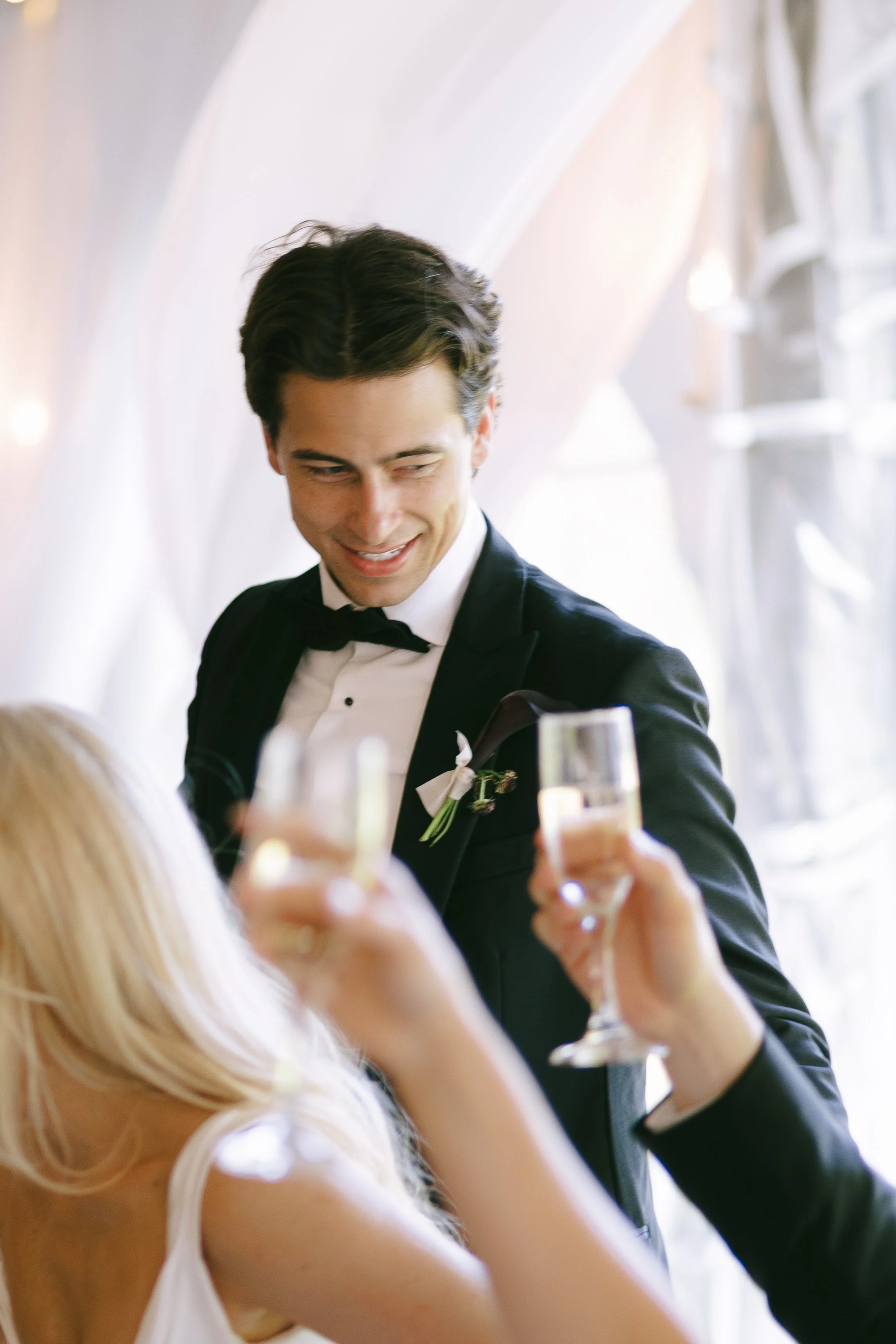 A man in a tuxedo and bow tie smiling and toasting with a woman at a celebration or wedding.