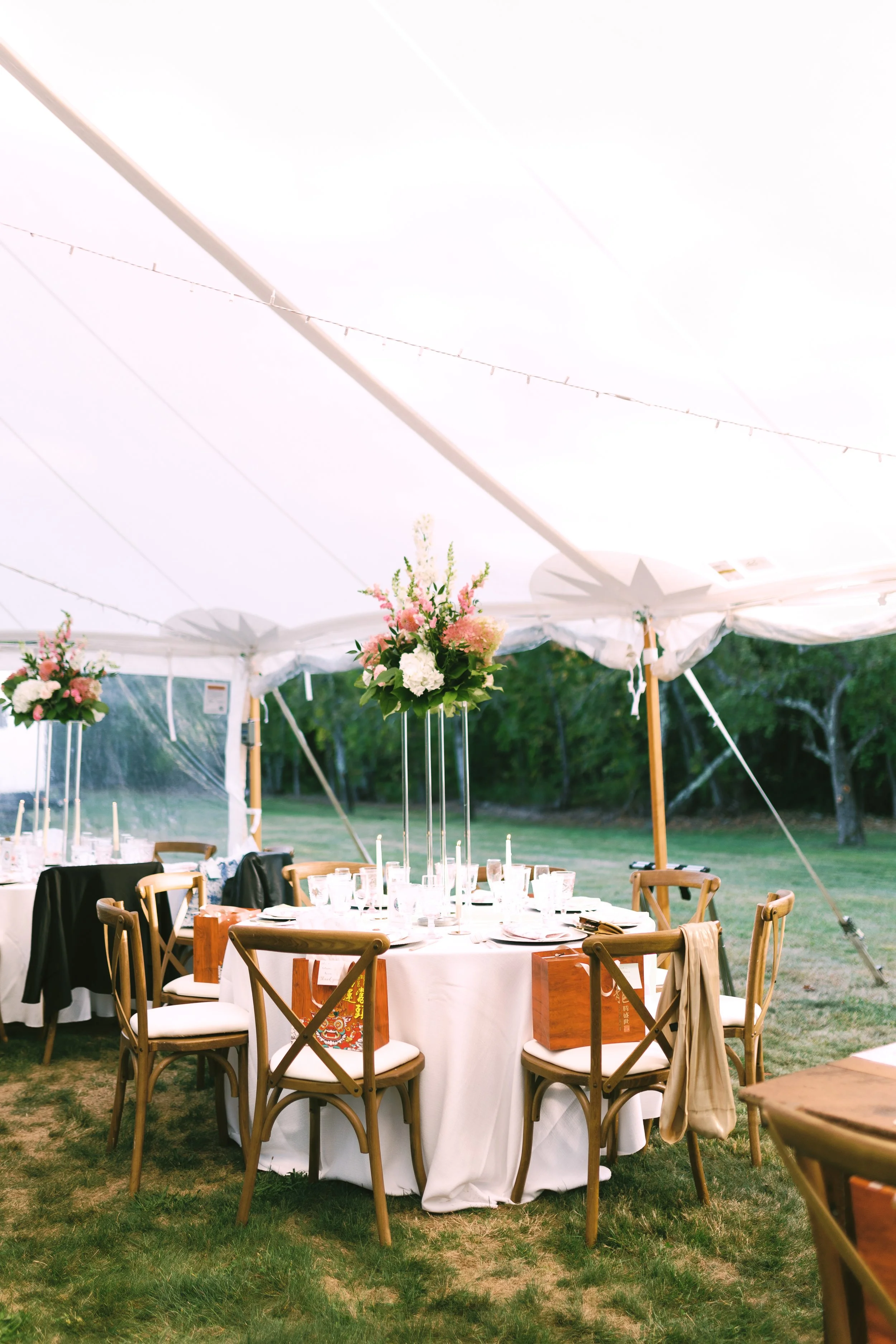 Wedding reception setup under a white tent with a round table decorated with pink and white flower centerpieces, surrounded by wooden chairs, on a grassy outdoor area.