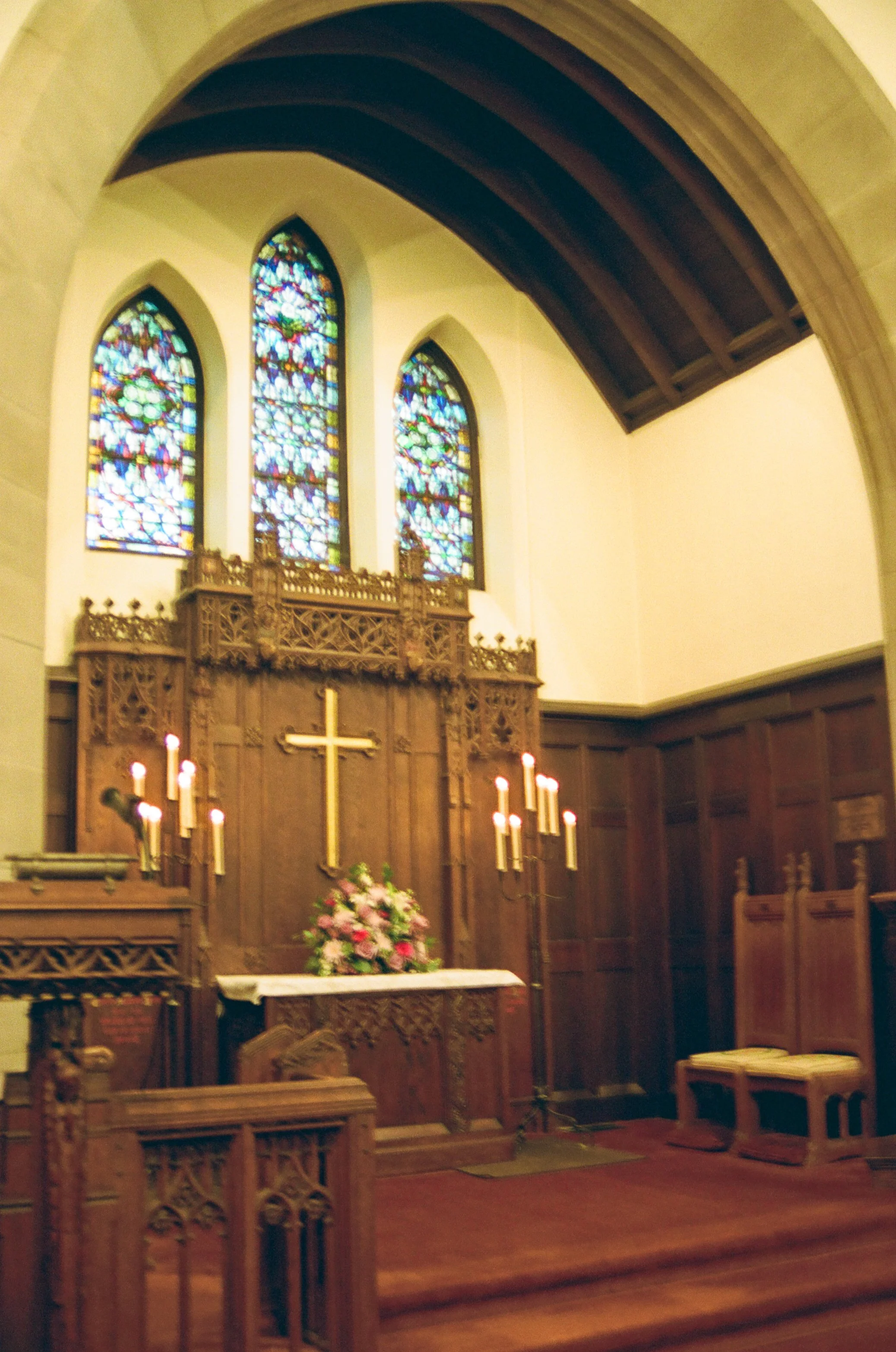 Inside a church with stained glass windows, an altar with a cross, flowers, and lit candles.