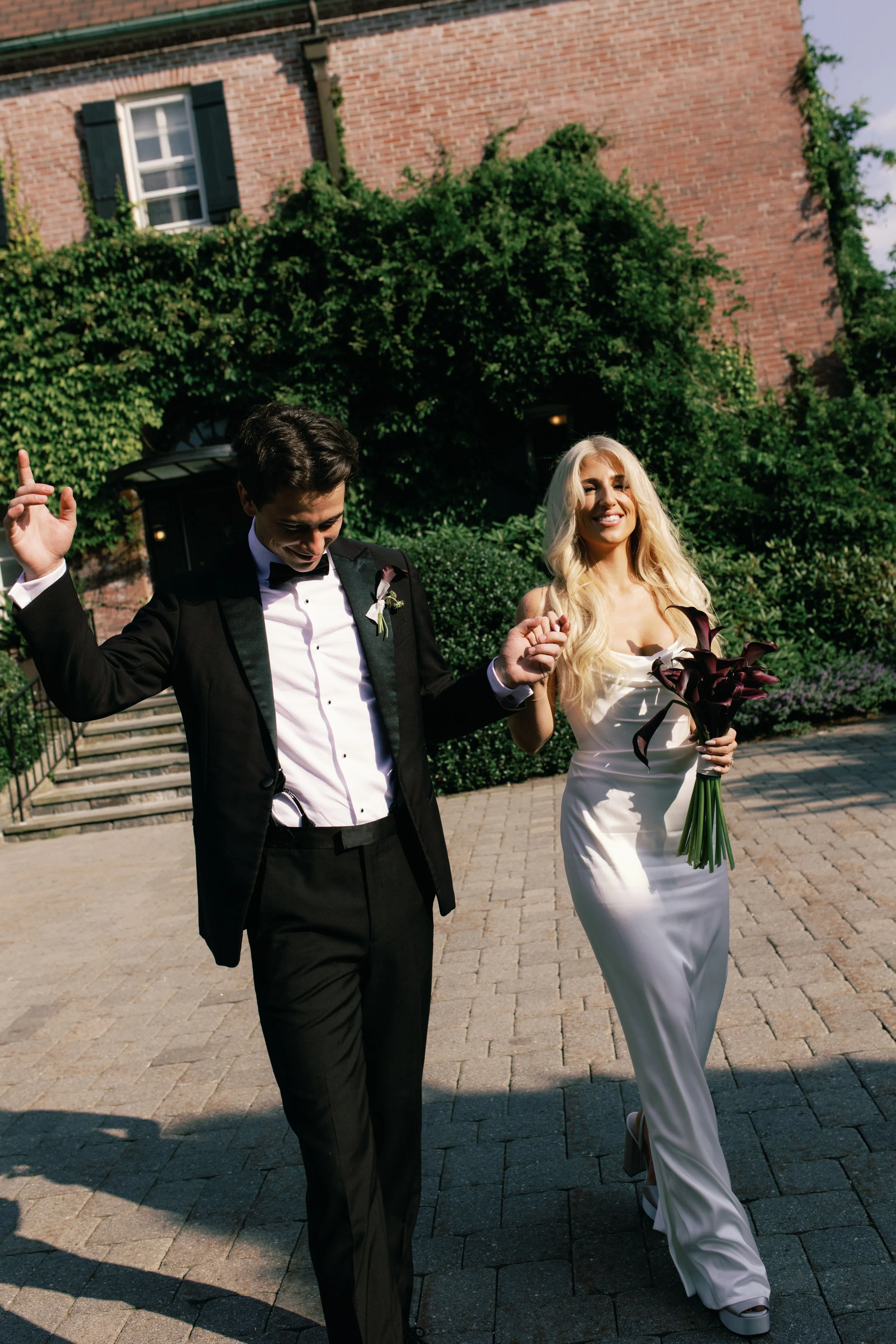 A bride in a white satin gown holding a bouquet of dark calla lilies, and a groom in a black tuxedo with a white shirt and black bowtie, walking outdoors on a paved path. They are holding hands, smiling, and appear to be celebrating their wedding.
