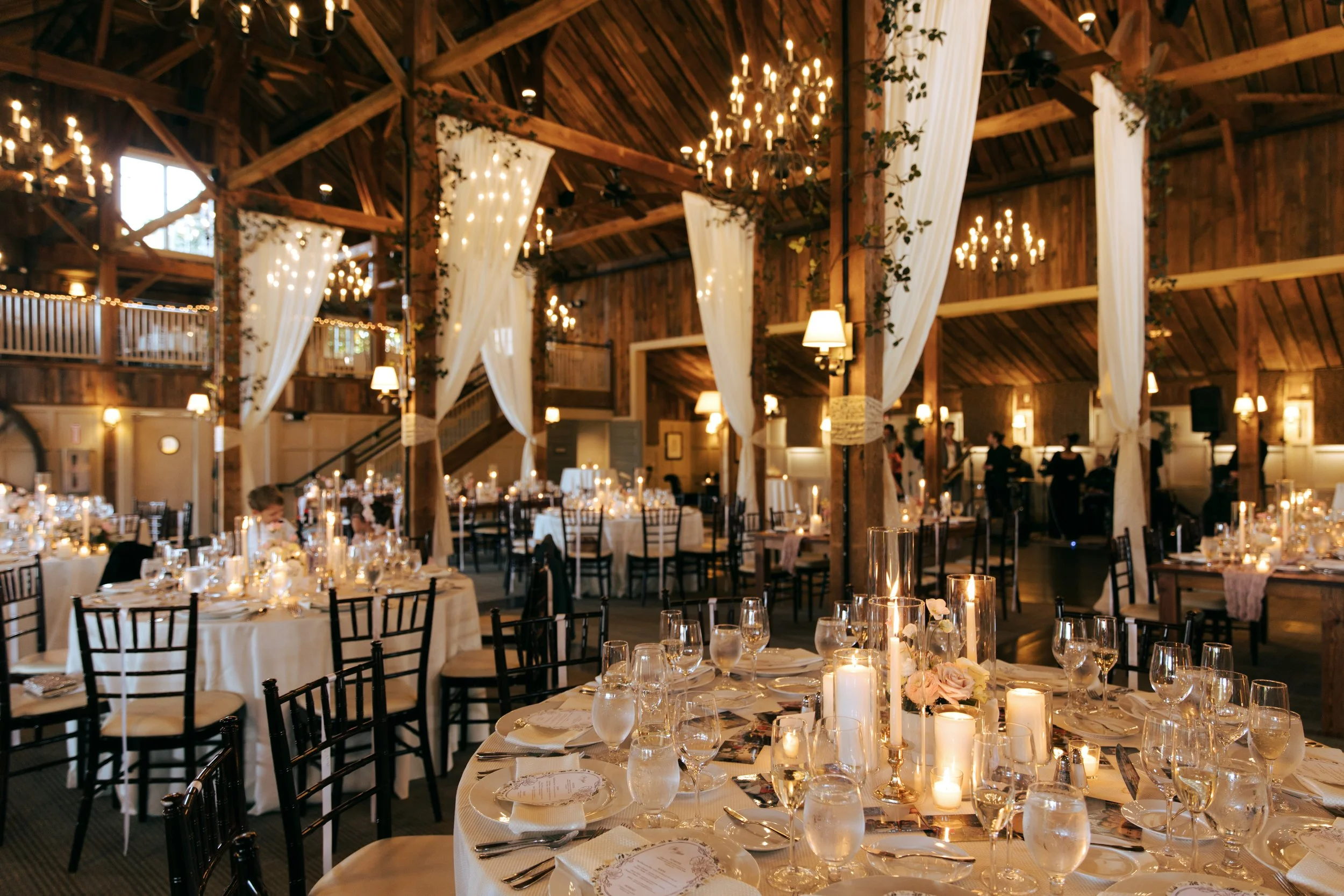 Barn at Gibbet Hill Groton Elegant wedding reception inside a rustic wooden barn, decorated with chandeliers, white drapes, and string lights, featuring round tables set with candles, flowers, glassware, and black chairs.