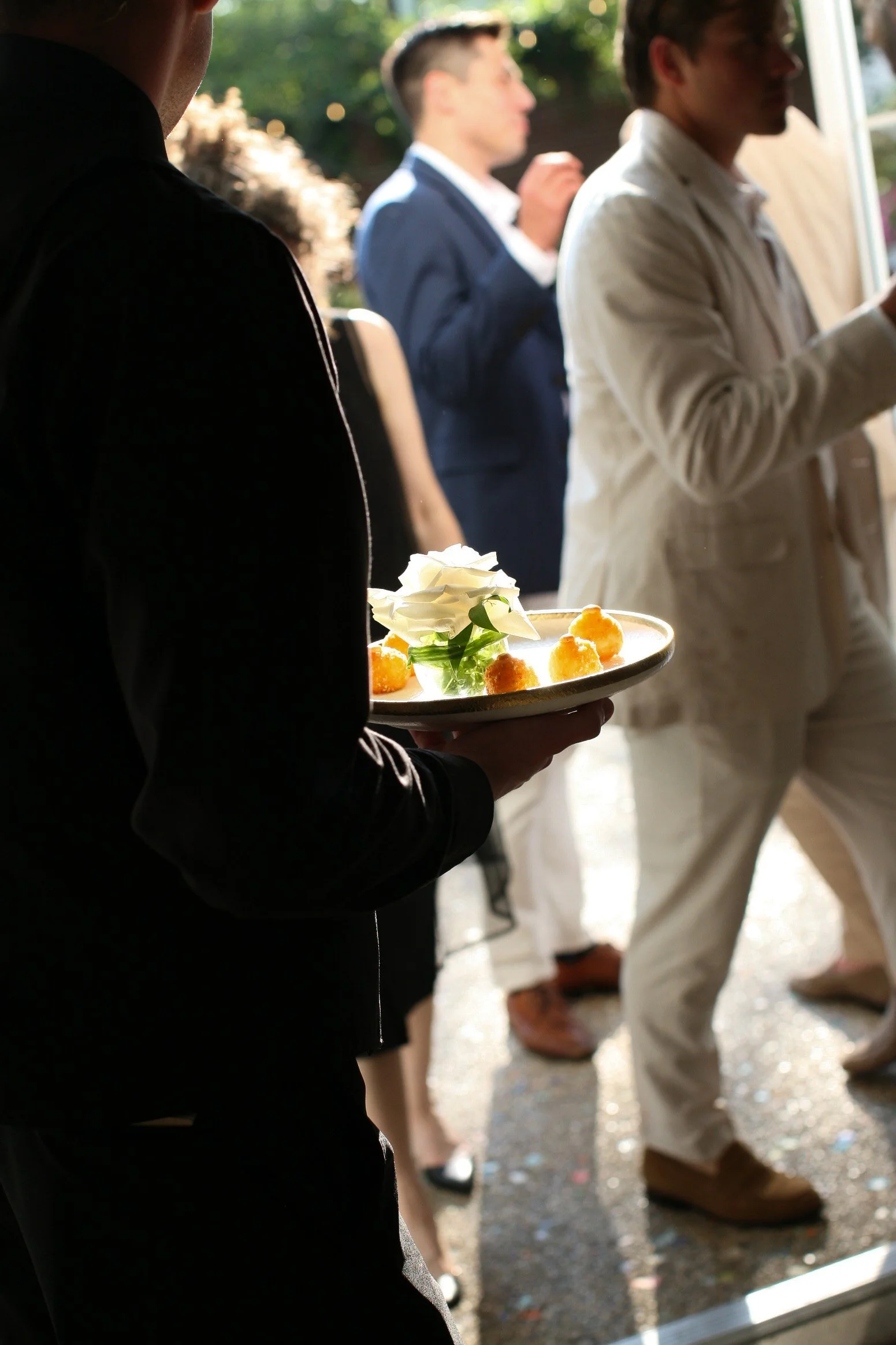 A person in black holding a plate with white flowers and small breaded appetizers at a gathering or party.