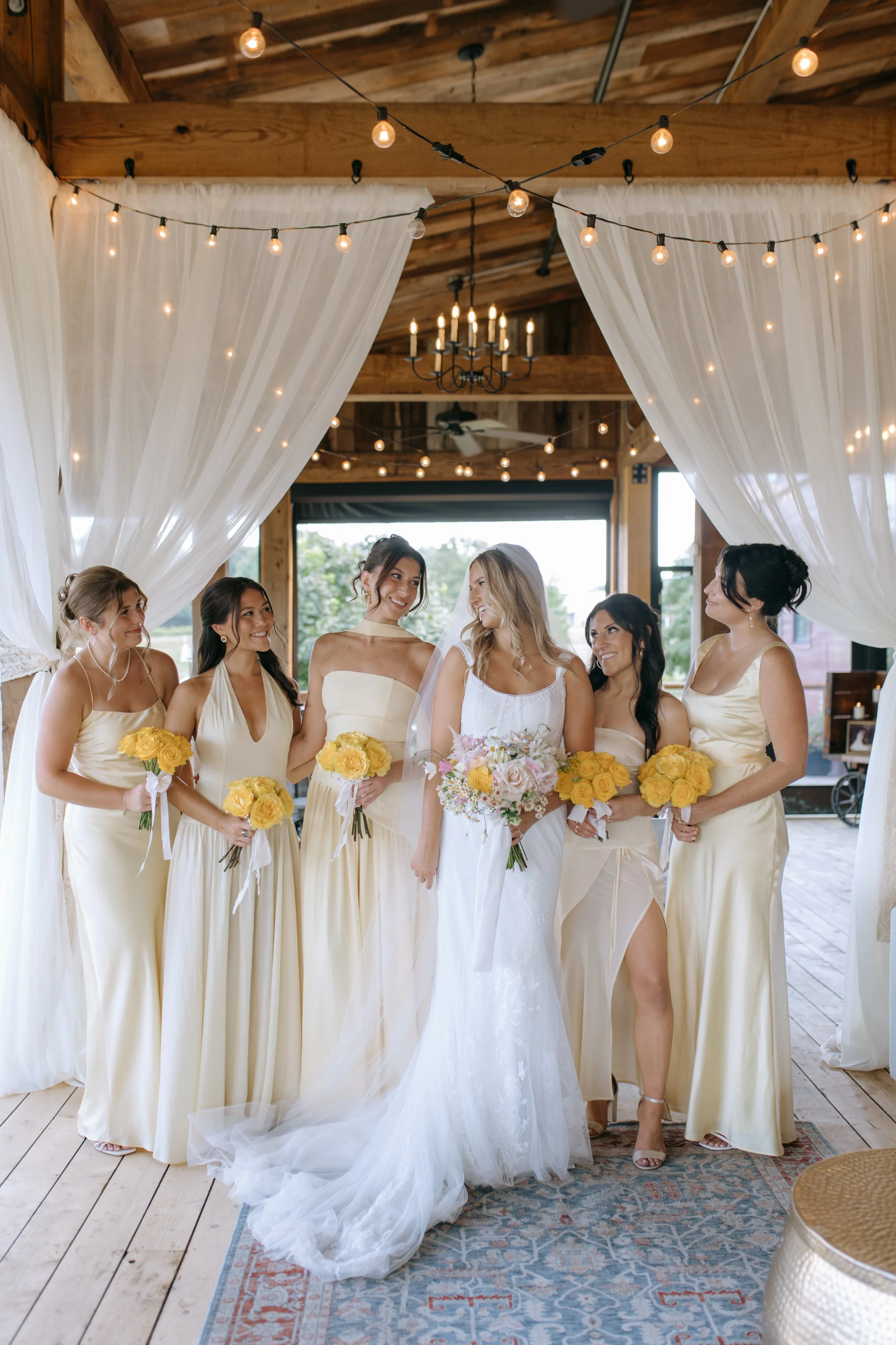 A group of seven women, including a bride and her bridesmaids, standing inside a decorated venue with draped white curtains and string lights, holding bouquets of yellow roses. The bride wears a white wedding gown, and the bridesmaids wear matching c