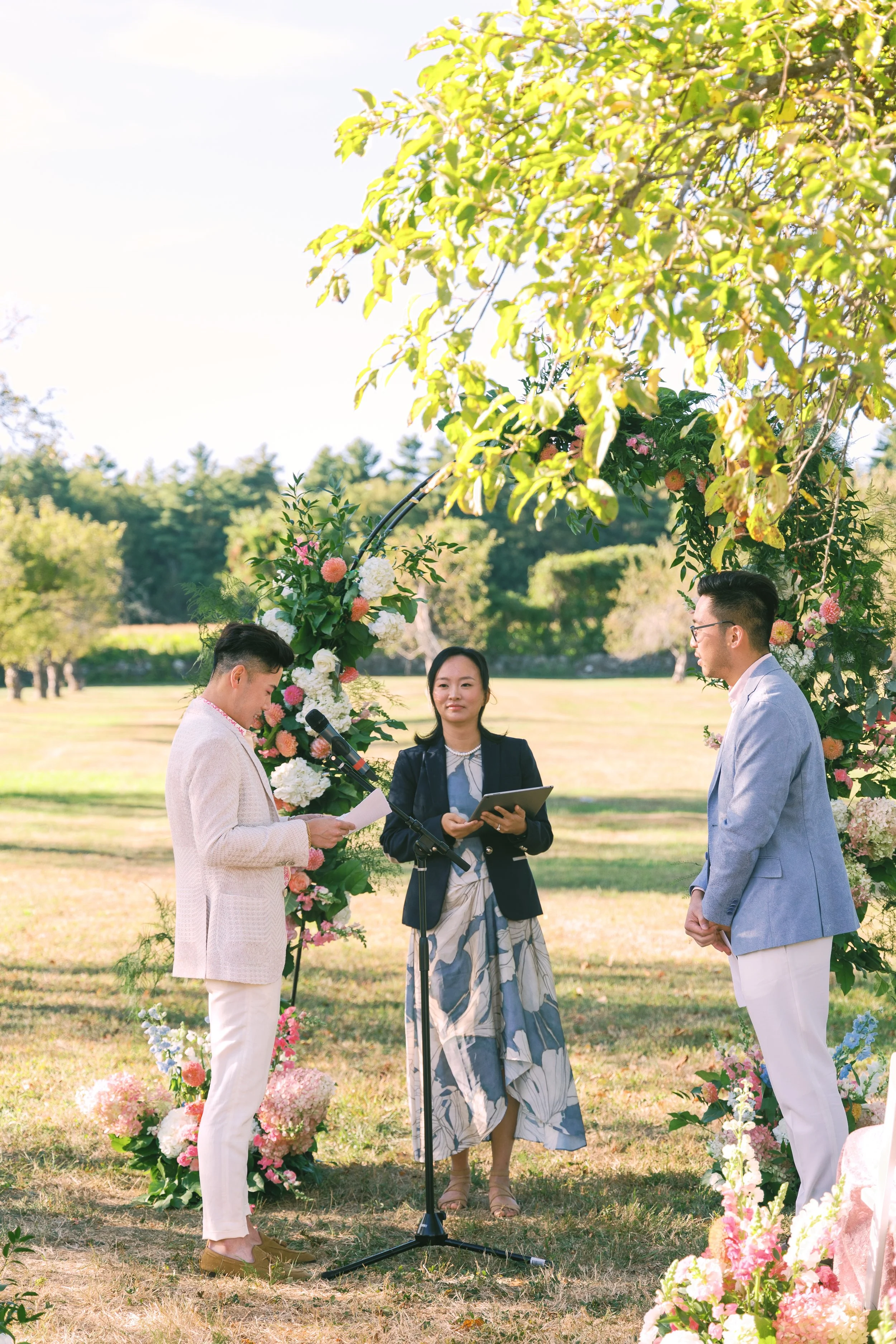 A wedding ceremony outdoors with a couple standing under a floral arch, with a woman officiating and reading from a paper, on a sunny day surrounded by trees and greenery.