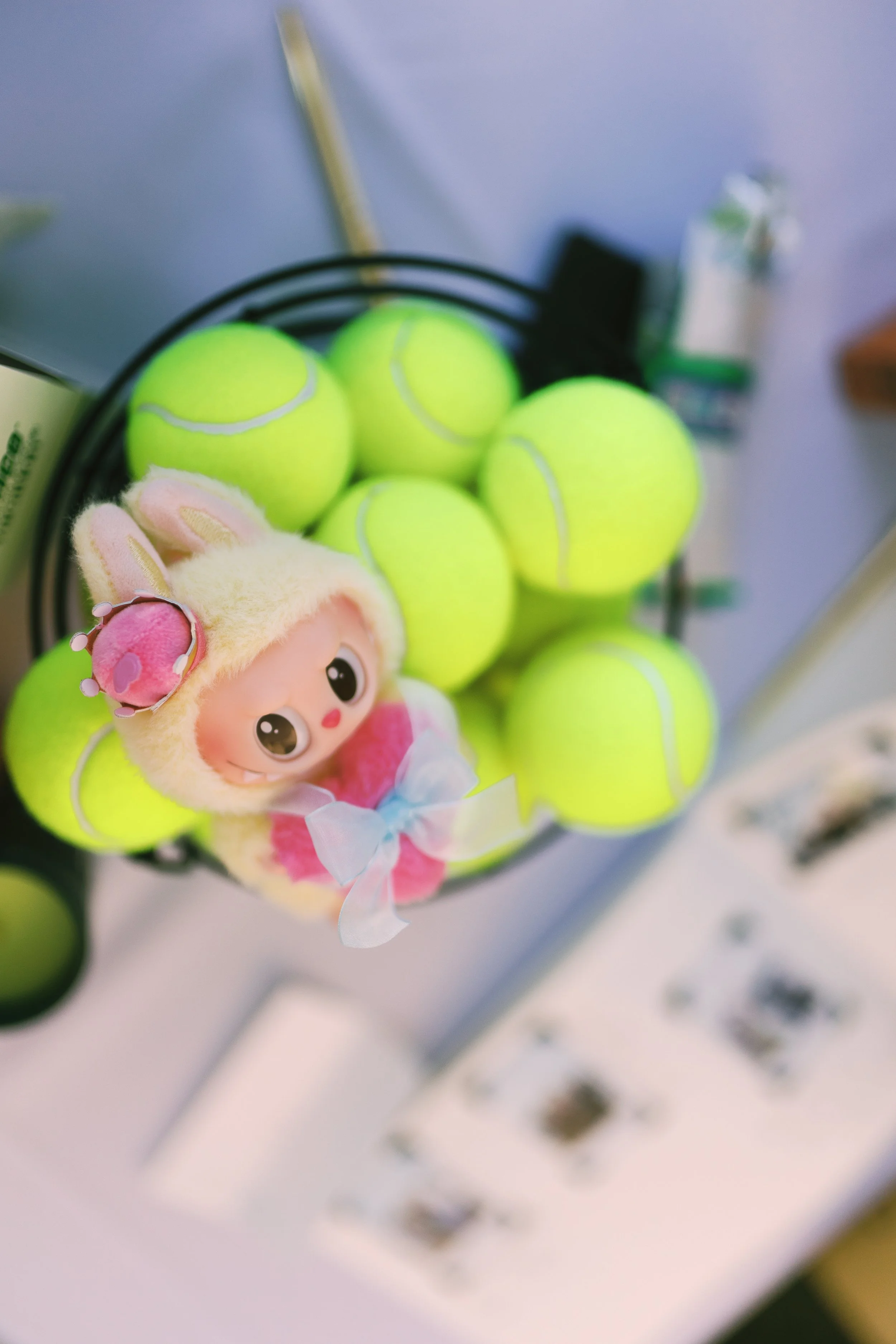A basket of bright yellow tennis balls, with a pink and white plush toy with bunny ears and a blue bow on top.