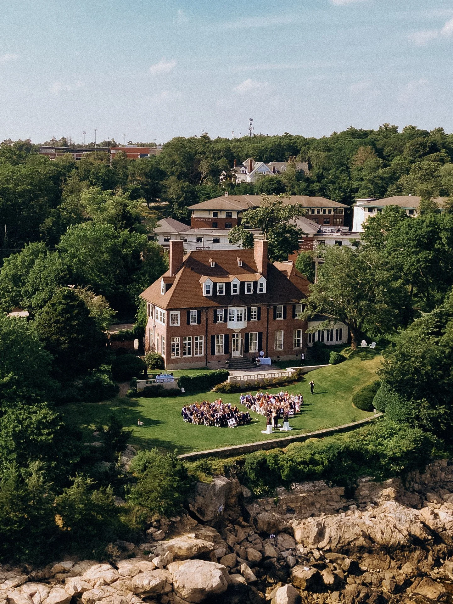 A wedding ceremony taking place on the lawn in front of a large brick house surrounded by trees.